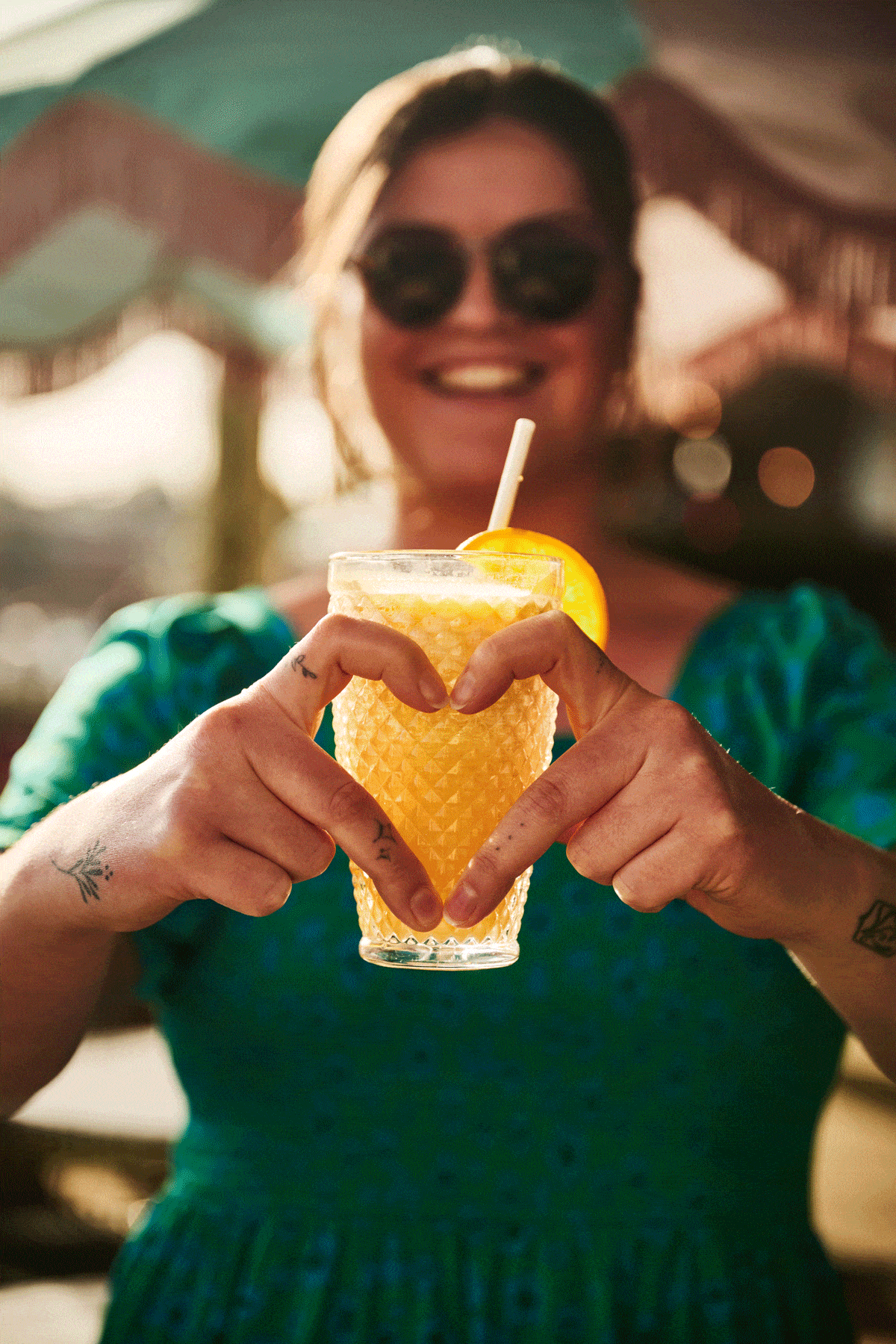 A smiling woman in sunglasses and a green dress holds a yellow drink with a lemon slice, forming a heart shape with her hands around the glass. The sunny, blurred background hints at the vibrant atmosphere of Torino.