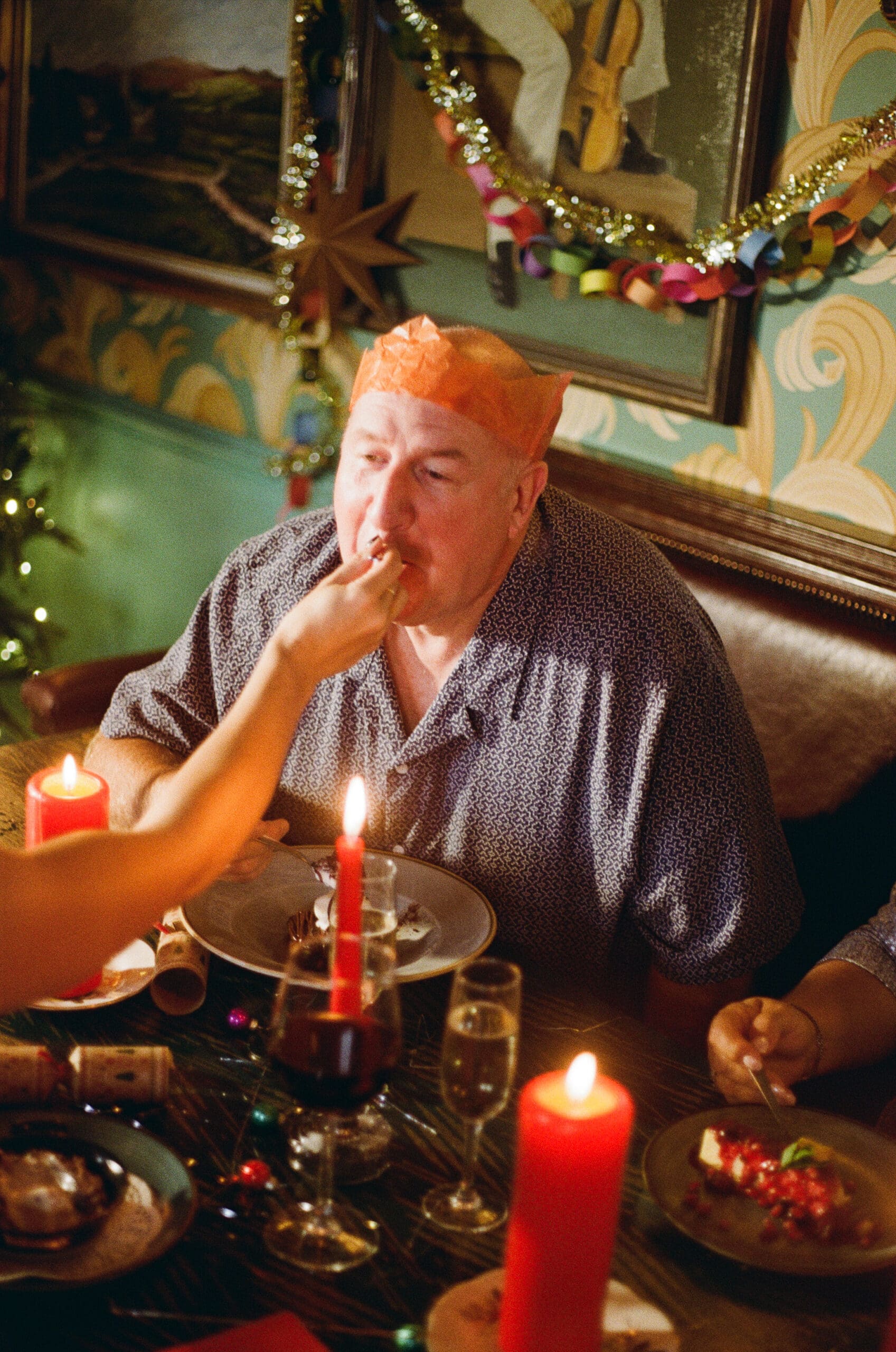 A person wearing a paper crown sits at a festive Christmas dinner table while someone else feeds them. The table is decorated with candles, drinks, and holiday decorations, with garlands and art on the wall in the background.