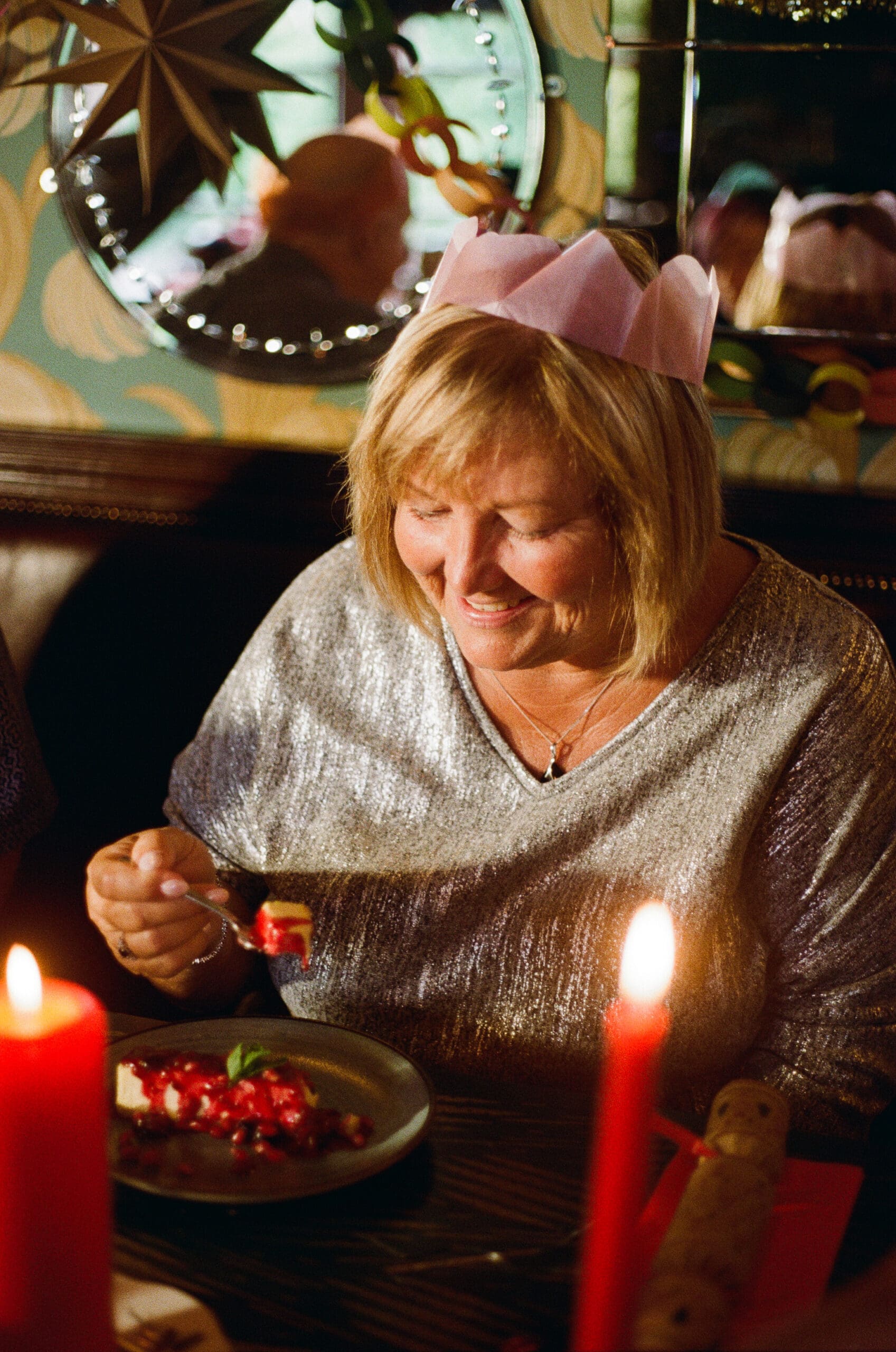 A smiling woman wearing a pink paper crown and a sparkly top sits at a table with red candles, holding a fork and enjoying a dessert topped with berries. The setting appears festive and warm.