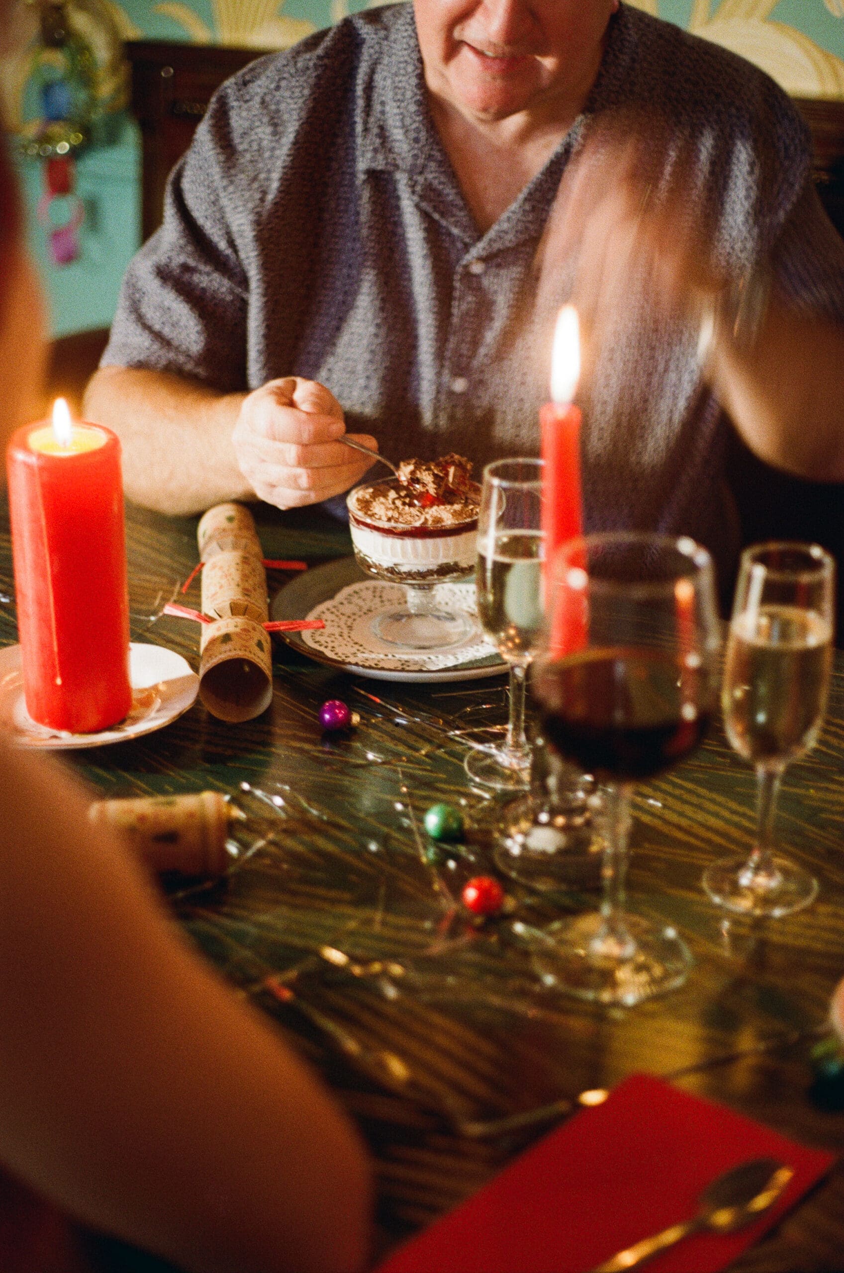 A person sits at a festive table with candles, wine glasses, and holiday decorations, holding a spoon over a dessert. Another person is partially visible in the foreground.
