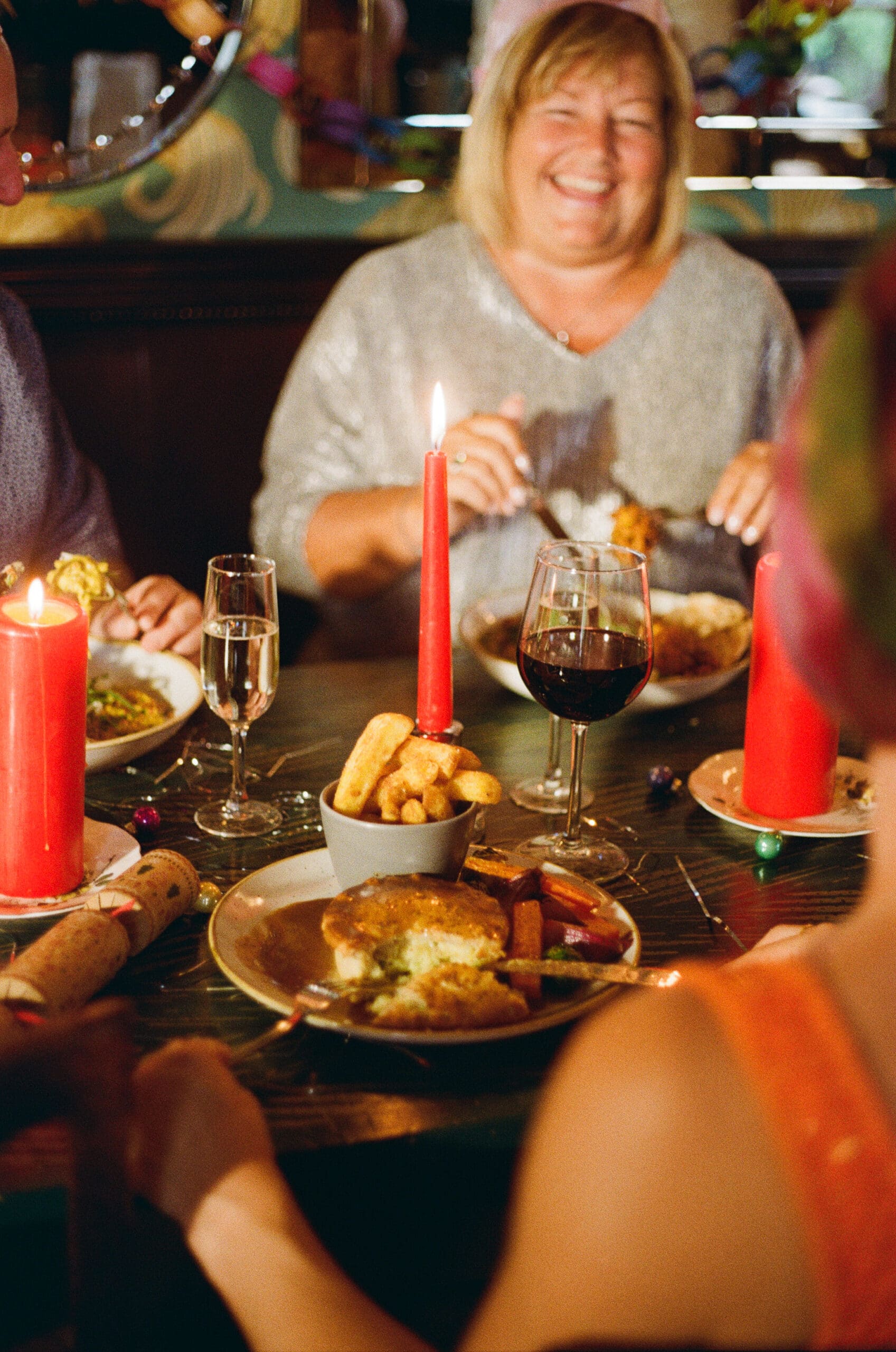 A smiling person sits at a candle-lit table with others, enjoying a festive Christmas meal. Plates of food, wine, champagne, and red candles fill the wooden table, creating a warm, celebratory atmosphere.
