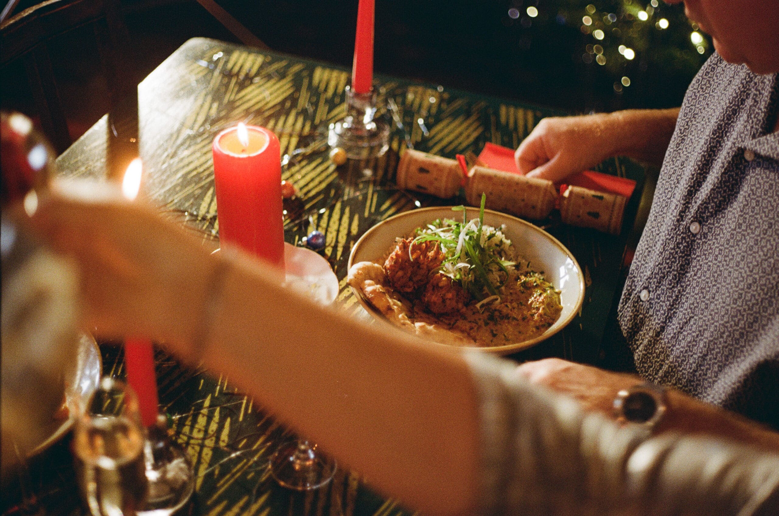 A person sits at a festive table with a plate of food, lit red candles, and a Christmas cracker. Another hand reaches across the table, and there are decorations suggesting a holiday meal.