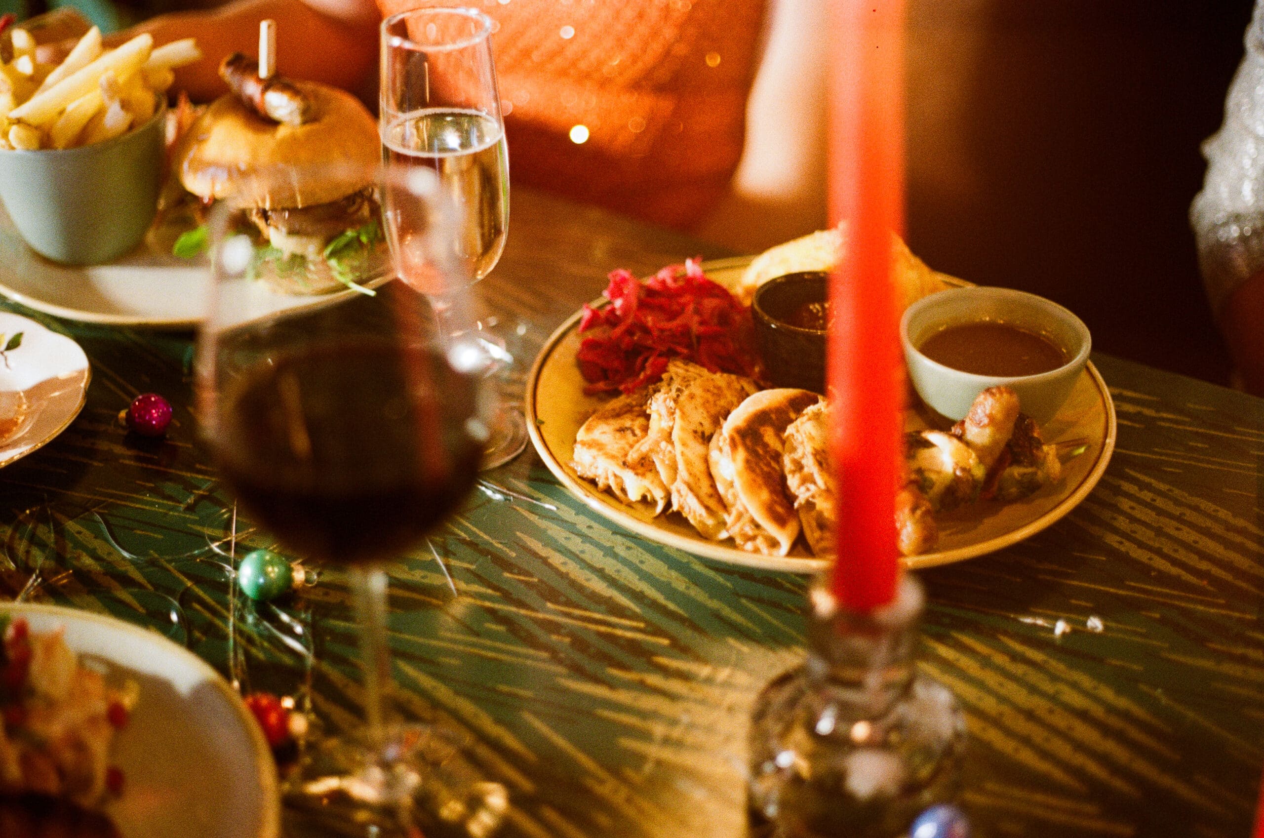 A festive dining table set with plates of food, including a burger with fries and a platter of sliced roast with sides, a glass of red wine, champagne, and a red candle in the foreground.