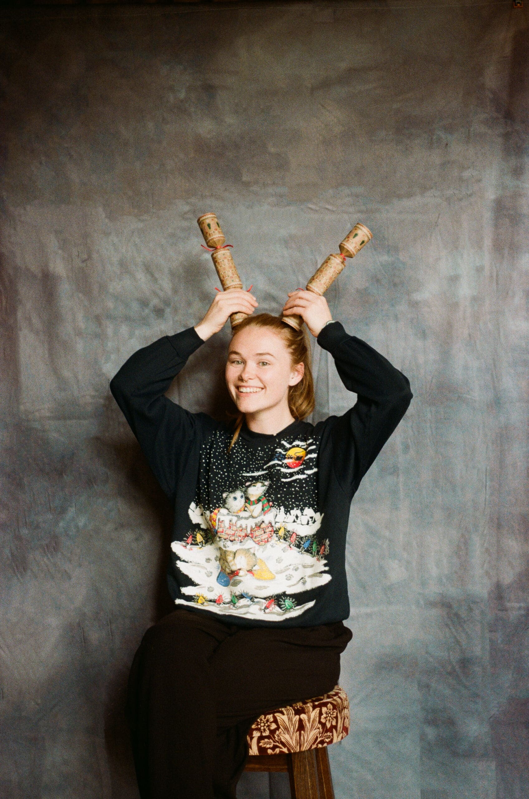 A smiling person in a festive Christmas sweater with a winter animal scene sits on a stool, holding two wooden objects above their head like antlers against a neutral backdrop.