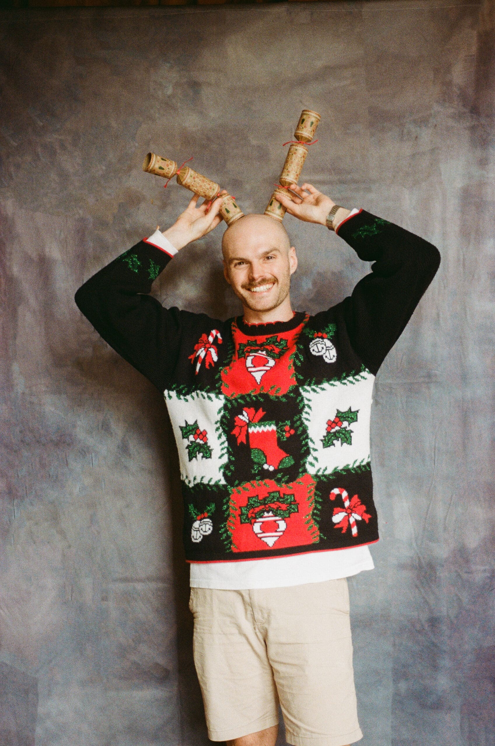 A smiling person in a festive Christmas sweater and shorts holds two Christmas crackers above their head like reindeer antlers, standing in front of a gray backdrop.
