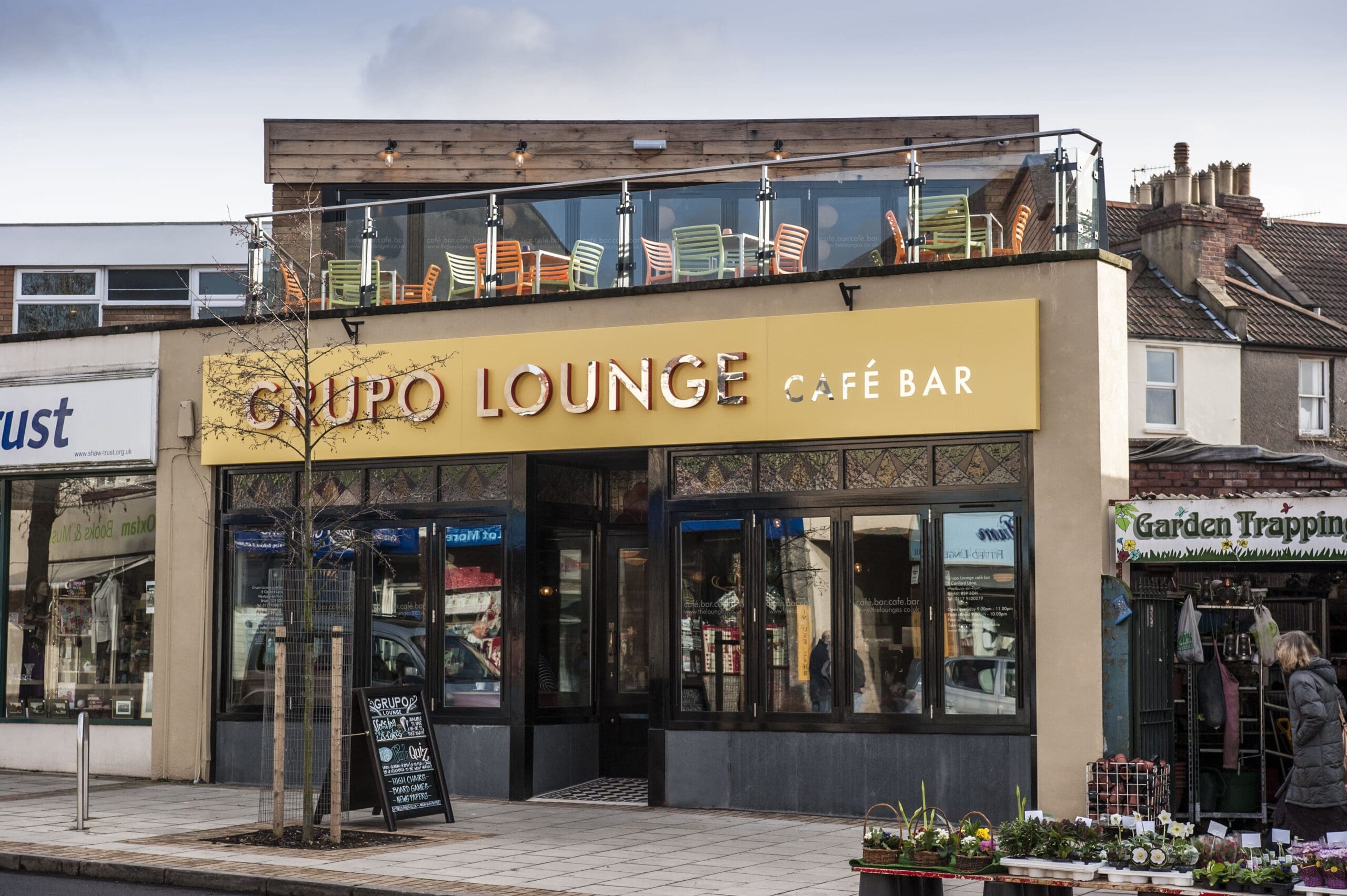 A street view of Grupo Lounge Café Bar with a yellow Grupo sign, large windows, and a rooftop terrace adorned with colorful chairs. Nearby, shops line the street and pedestrians stroll past this inviting café bar.