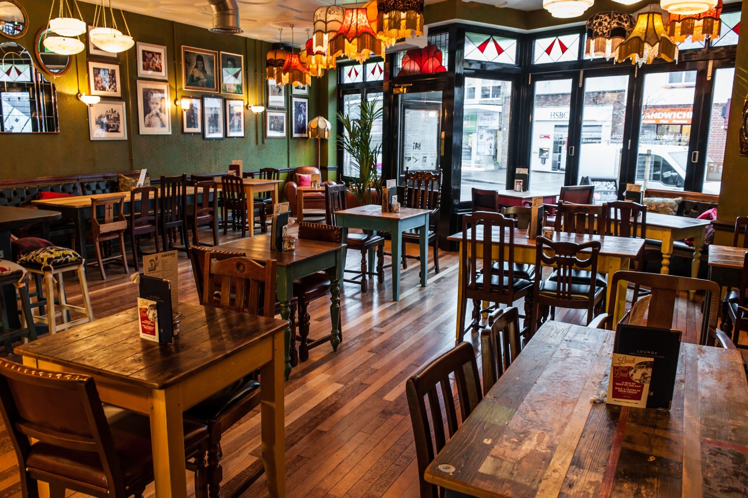 A cozy, eclectic café interior by Grupo with mismatched wooden tables and chairs, colorful hanging lamps, framed artwork on green walls, and large windows letting in natural light.