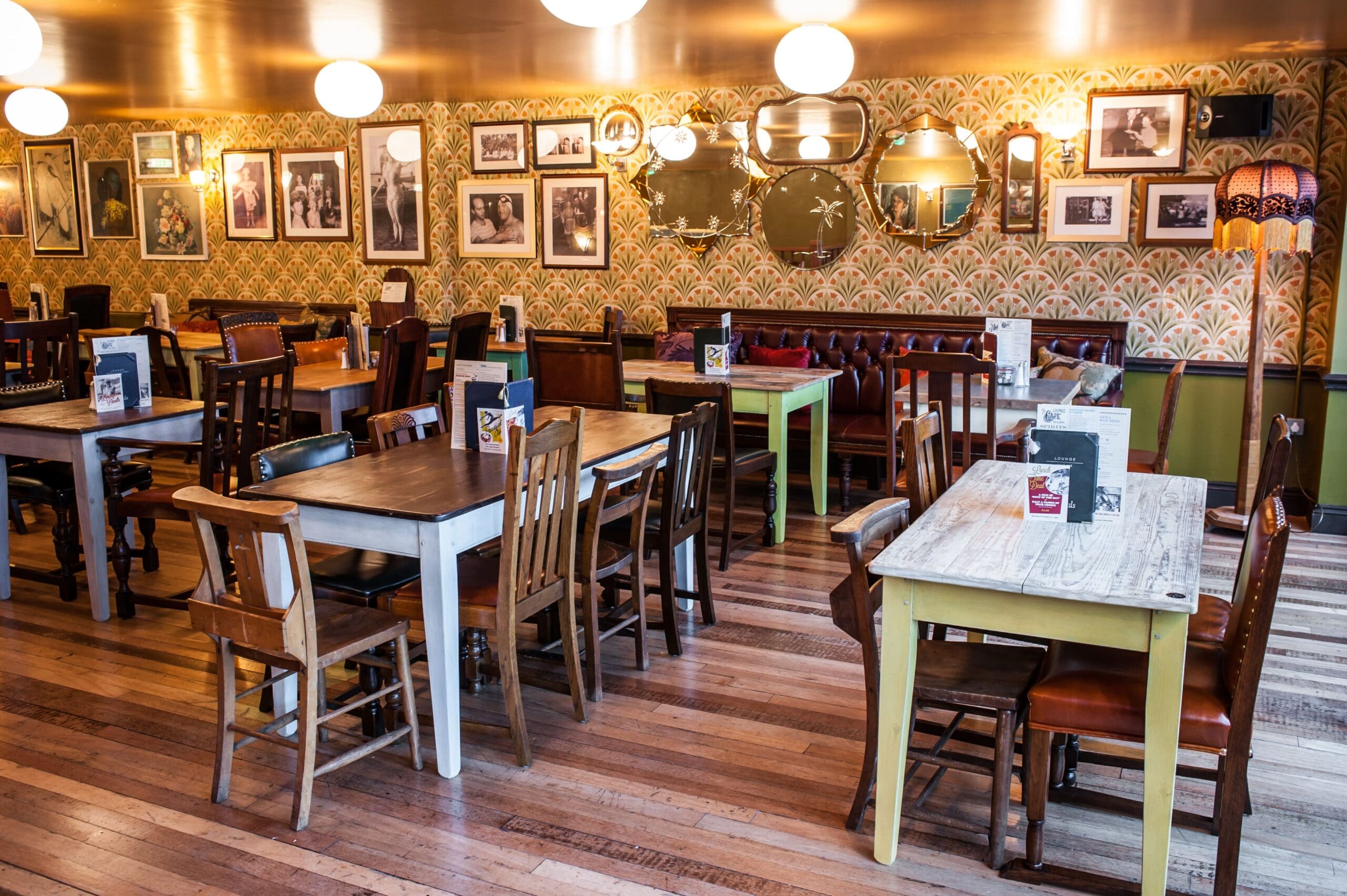 A cozy cafe by Grupo with mismatched wooden tables and chairs, patterned wallpaper, framed photos, and mirrors on the walls. The space is warmly lit with round ceiling lights and a vintage floor lamp.