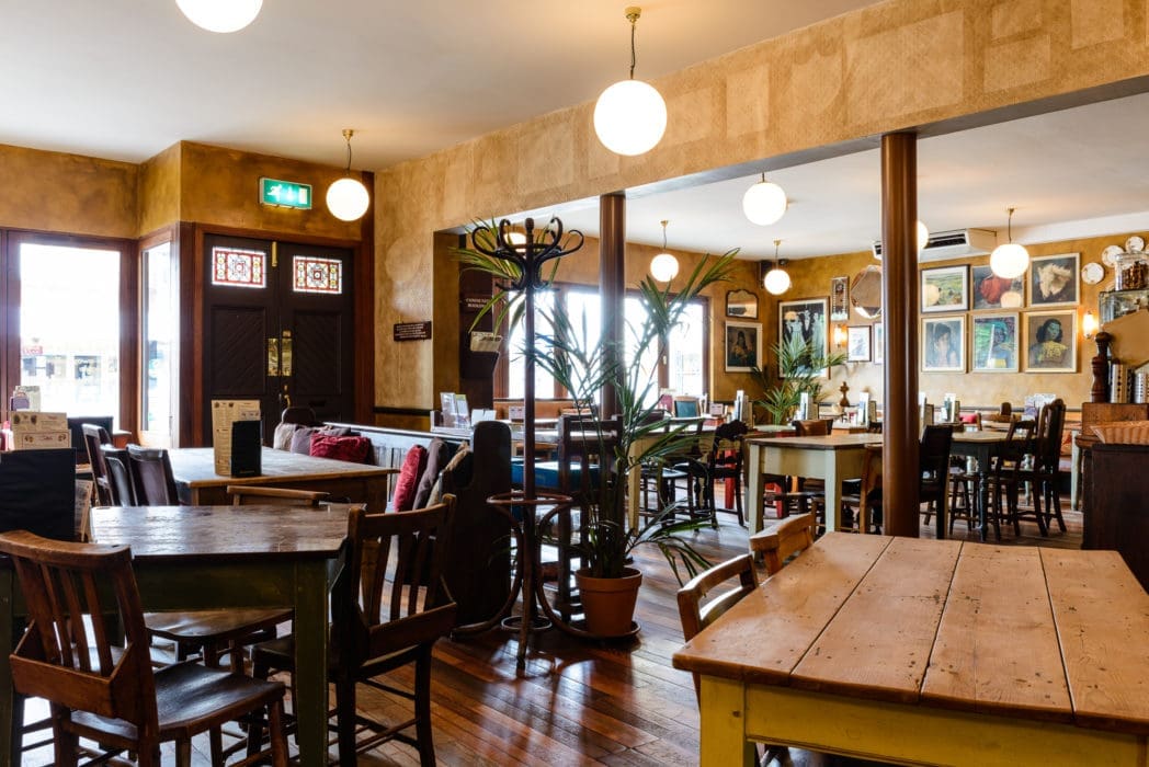 A cozy cafe interior with wooden tables and chairs, potted plants, round hanging lights, and framed artwork on the walls. Large windows fill Alto with light, enhancing its warm, inviting atmosphere.