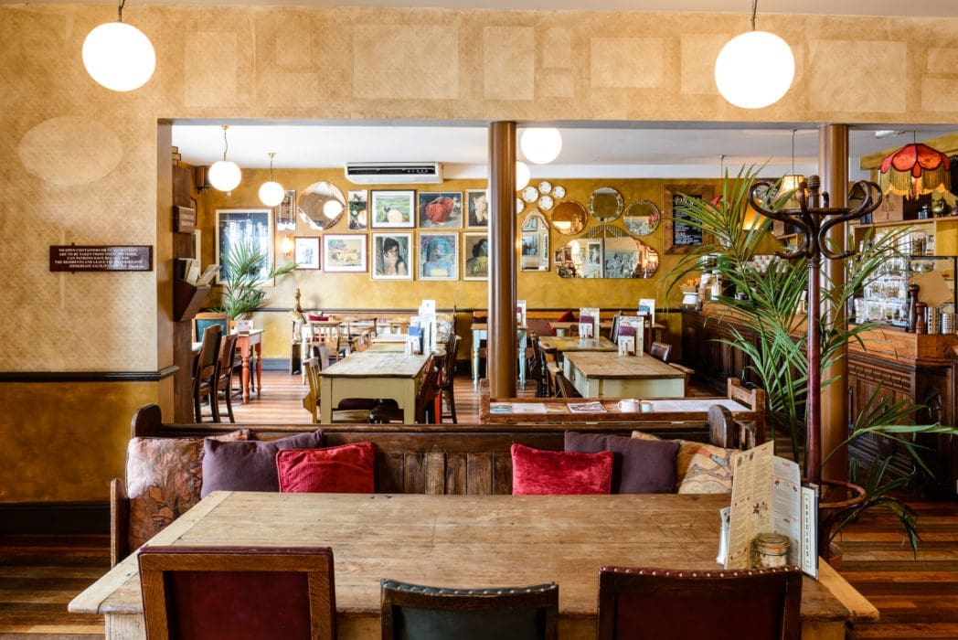 Cozy café with wooden tables and chairs, colorful cushions, framed art on yellow walls, hanging globe lights, plants, and vintage Alto decor style, creating a warm and inviting atmosphere.