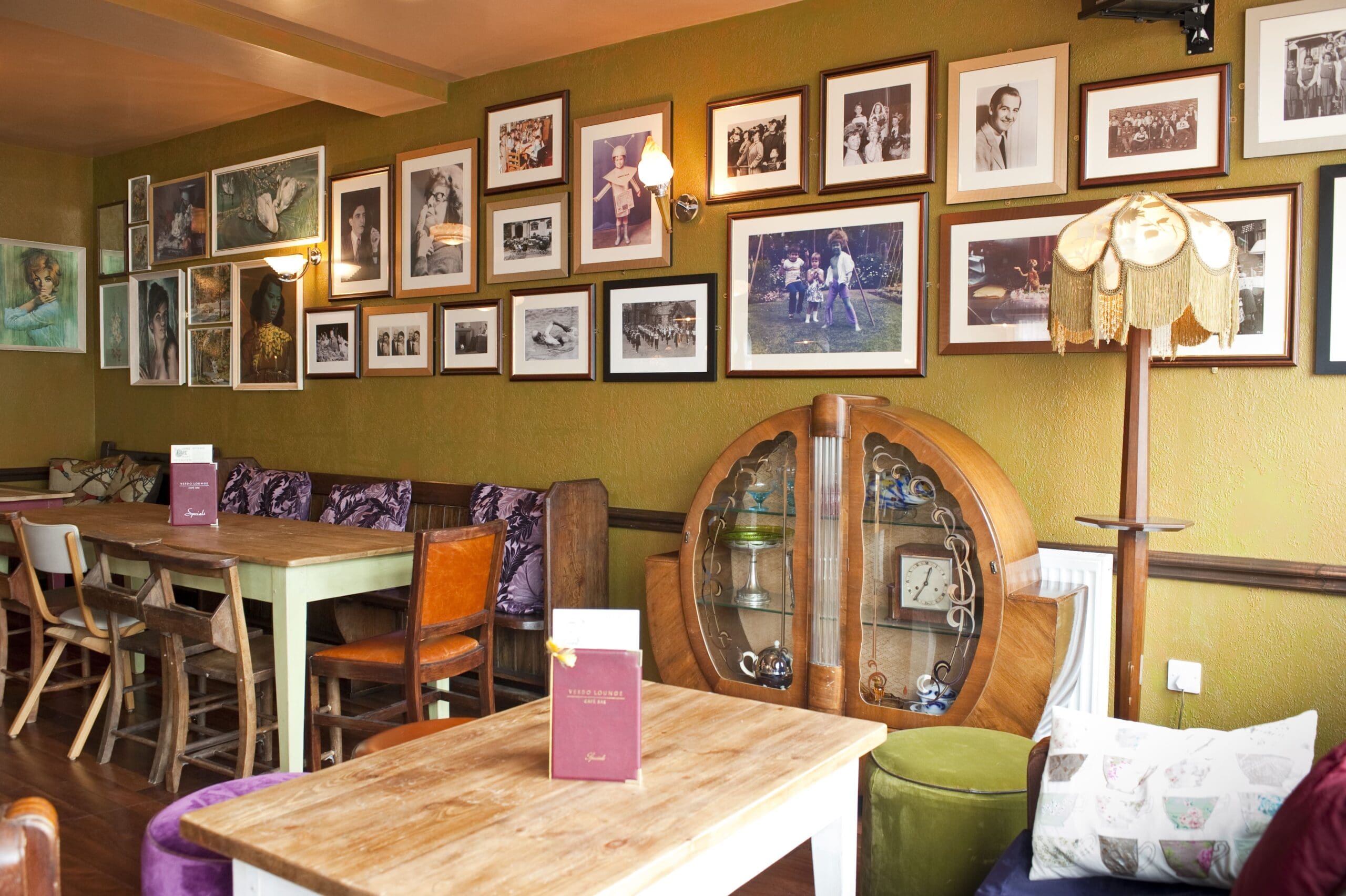 A cozy cafe with wooden tables and mismatched chairs, Verdo’s vintage decor shines with framed photos covering a green wall and a round glass cabinet beside a retro lampshade. Menus are placed on the tables.
