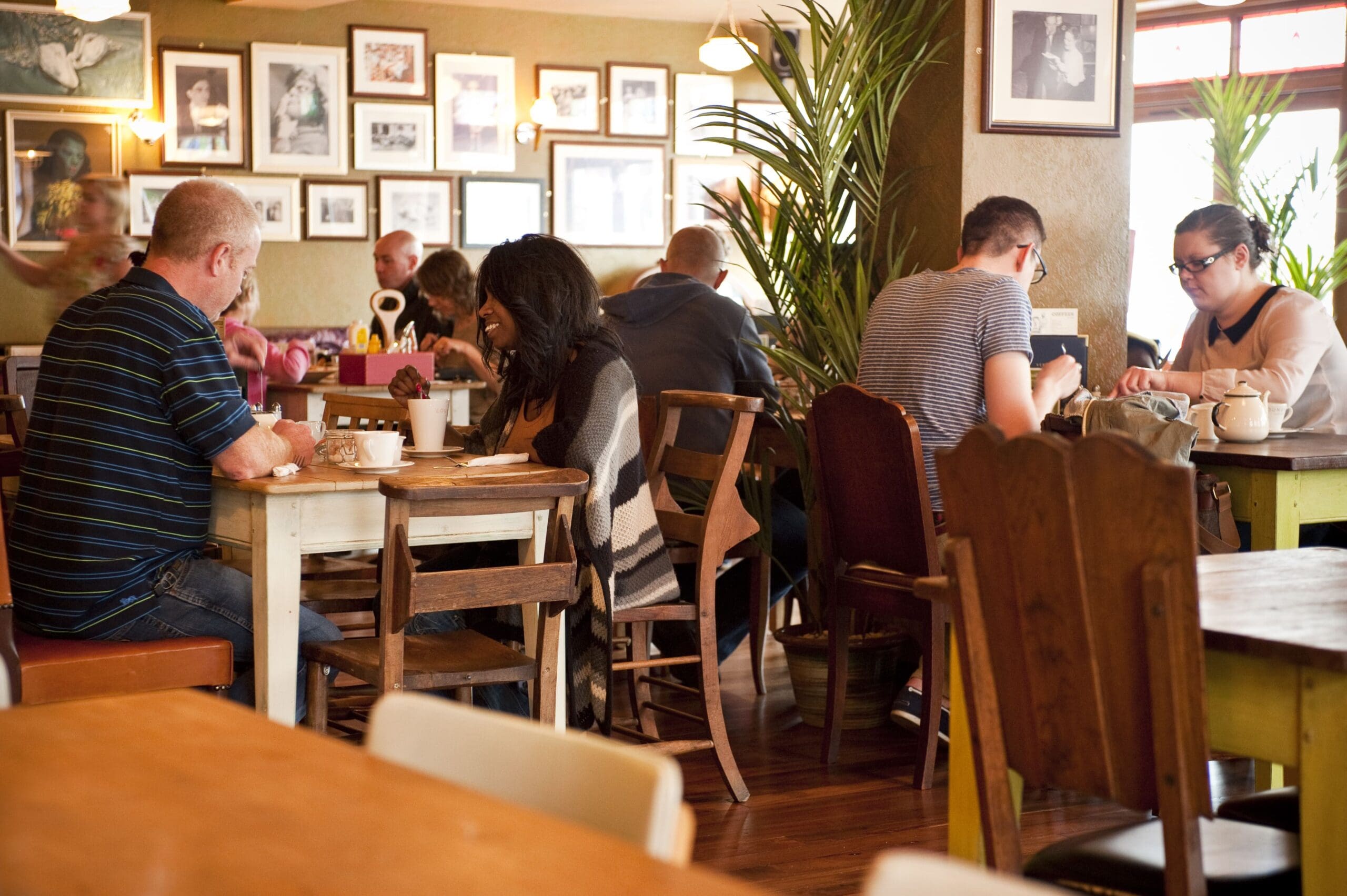 People sit at wooden tables in Verdo’s cozy café, eating, drinking, and talking. Framed photos decorate the walls, and plants add greenery to the warmly lit, relaxed atmosphere.