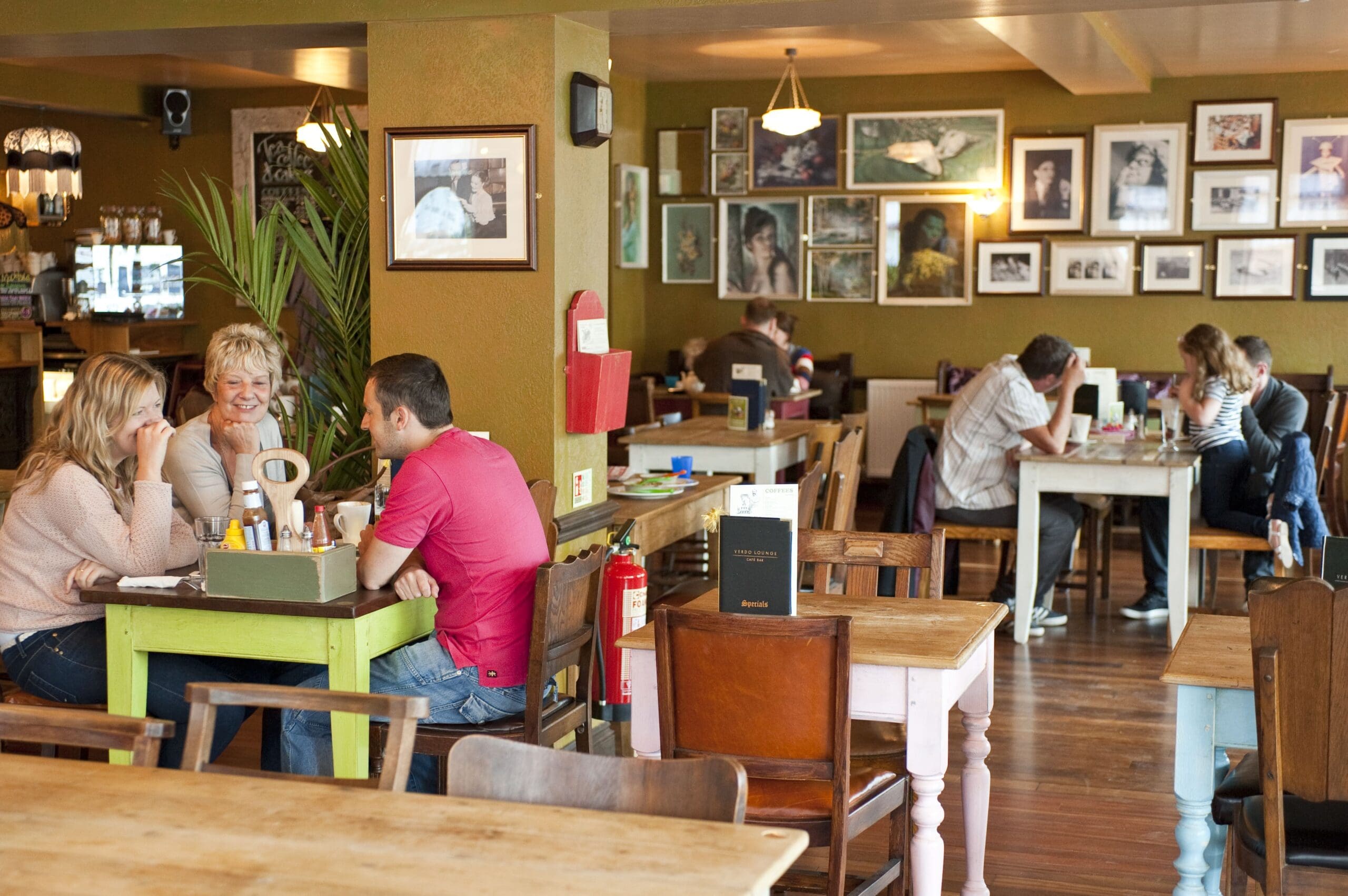 People sit and talk at wooden tables in Verdo, a cozy café with warm lighting. Framed photos decorate the olive-green walls, creating a relaxed atmosphere where groups chat and enjoy their time together.