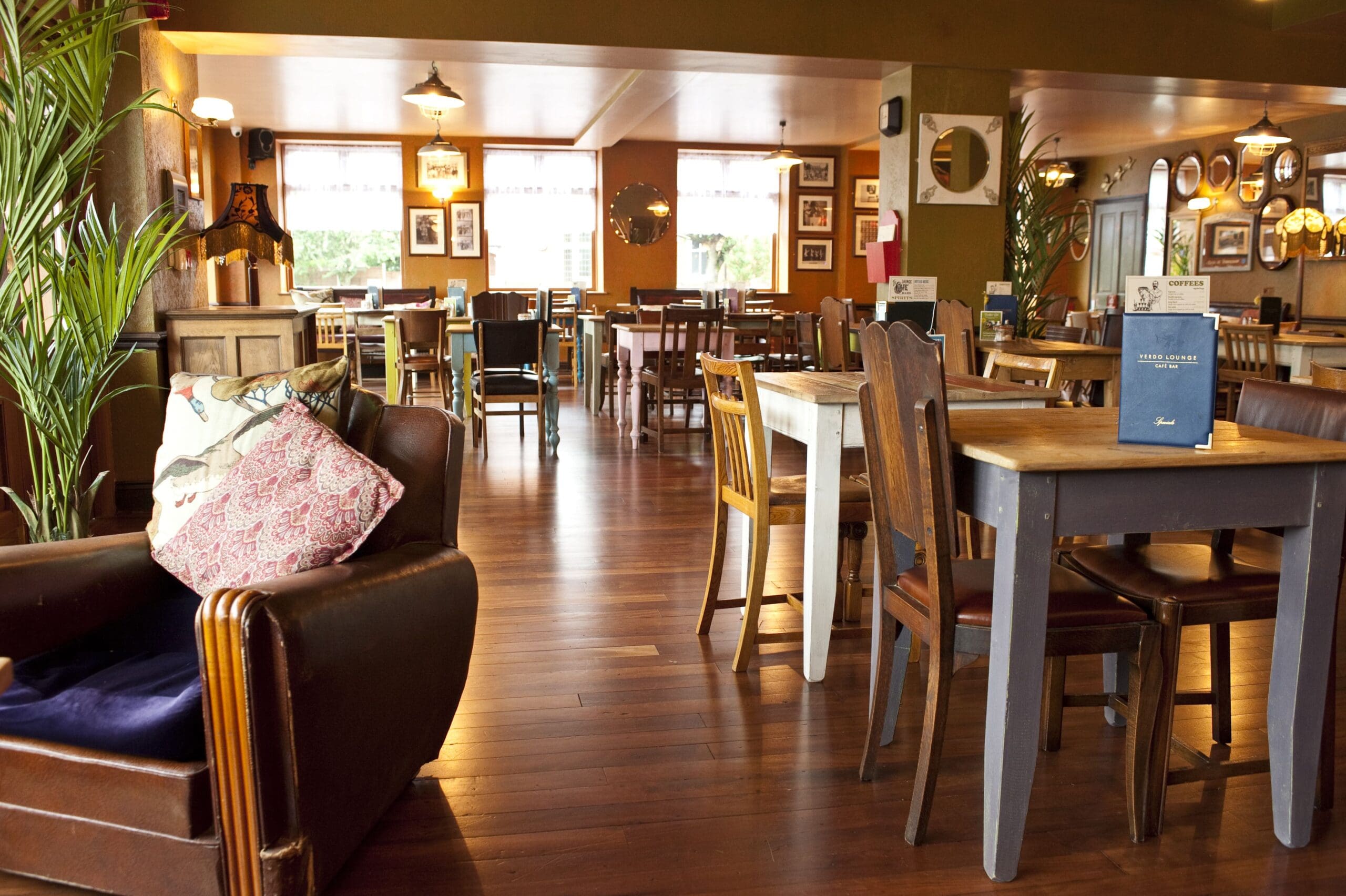 Cozy restaurant interior with wooden tables and chairs, soft lighting, green walls, and decorative mirrors. At Verdo, a brown leather armchair with patterned pillows sits among lush plants, creating a warm and inviting atmosphere.