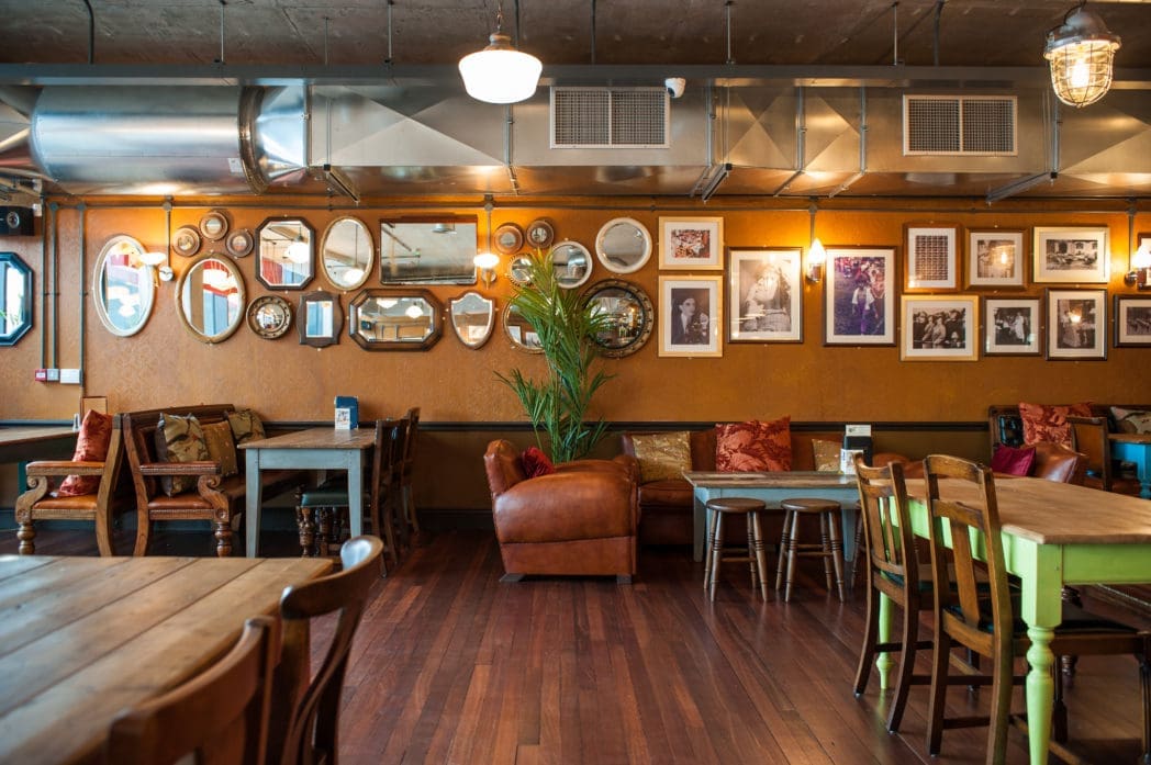 A cozy cafe by Brasco with wooden floors, assorted tables and chairs, cushioned seating, and a tan leather armchair. The orange wall is decorated with vintage mirrors, framed photos, and a large green plant.
