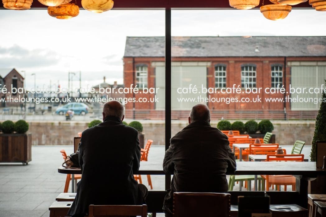 Two people sit at a table inside Brasco café, facing large windows overlooking a patio with orange chairs and tables, and a brick building in the background. Brasco branding is displayed on the windows.