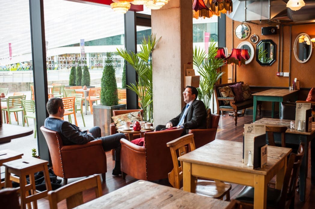 Two men in suits sit in armchairs having a conversation in a cozy, modern café with large windows, wooden tables, potted plants, and warm lighting—an inviting spot reminiscent of the signature Brasco atmosphere.