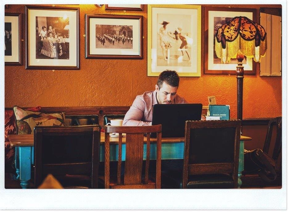 A man sits alone at a wooden table in Brasco, a cozy, warmly lit cafe, working on a laptop. Framed vintage photos and a decorative lamp adorn the orange wall behind him.