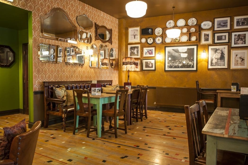 A cozy vintage café, Bosco, features wooden tables and chairs, patterned wallpaper, mirrors, framed photos, and plates adorning the gold-toned walls. Soft lighting creates a warm, inviting atmosphere.