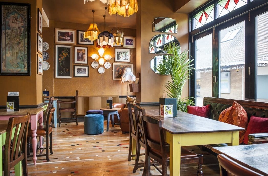 Cozy café interior with wooden tables, mixed chairs, colorful cushions, and art-filled walls. Large windows let in natural light, while plants and hints of Bosco green add a fresh touch to the inviting, eclectic space.