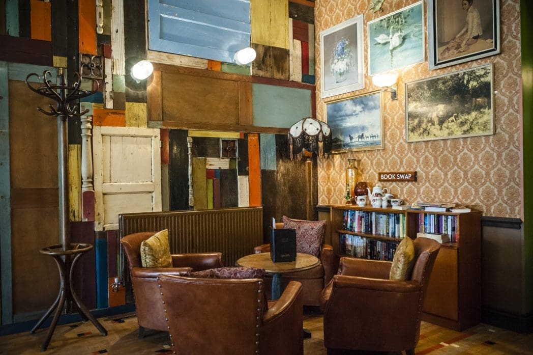 Cozy reading nook with three brown leather chairs around a small table, a bookshelf labeled “BOOK SWAP,” eclectic wall decor, vintage floor lamp, Bosco accents, and colorful patchwork wooden paneling.