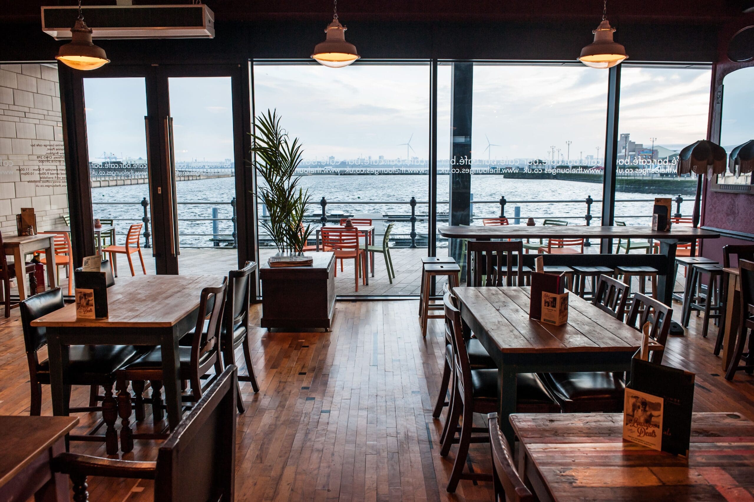 A cozy Marino cafe with wooden tables and chairs overlooks a waterfront through large glass windows. Outdoor seating is visible on the deck, and the scene is calm with distant wind turbines across the water.
