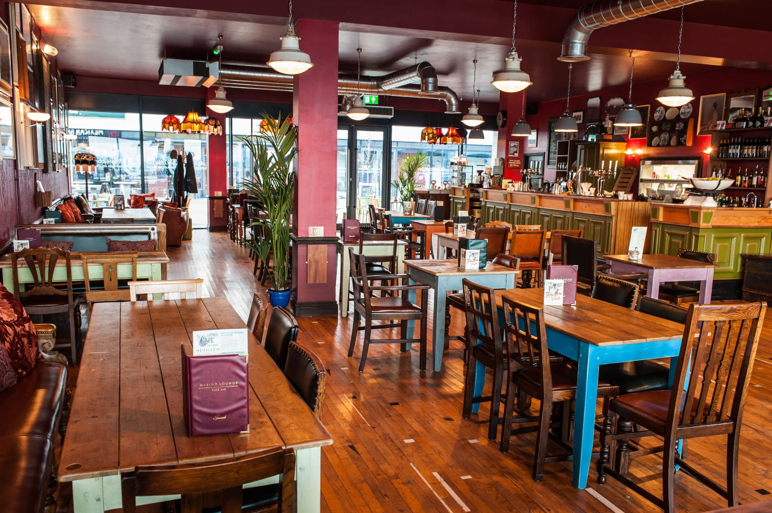 A colorful, cozy café or restaurant with wooden tables and mismatched Marino chairs, a long bar counter, indoor plants, and warm hanging lights creates a welcoming atmosphere. Menus and condiments are on the tables.