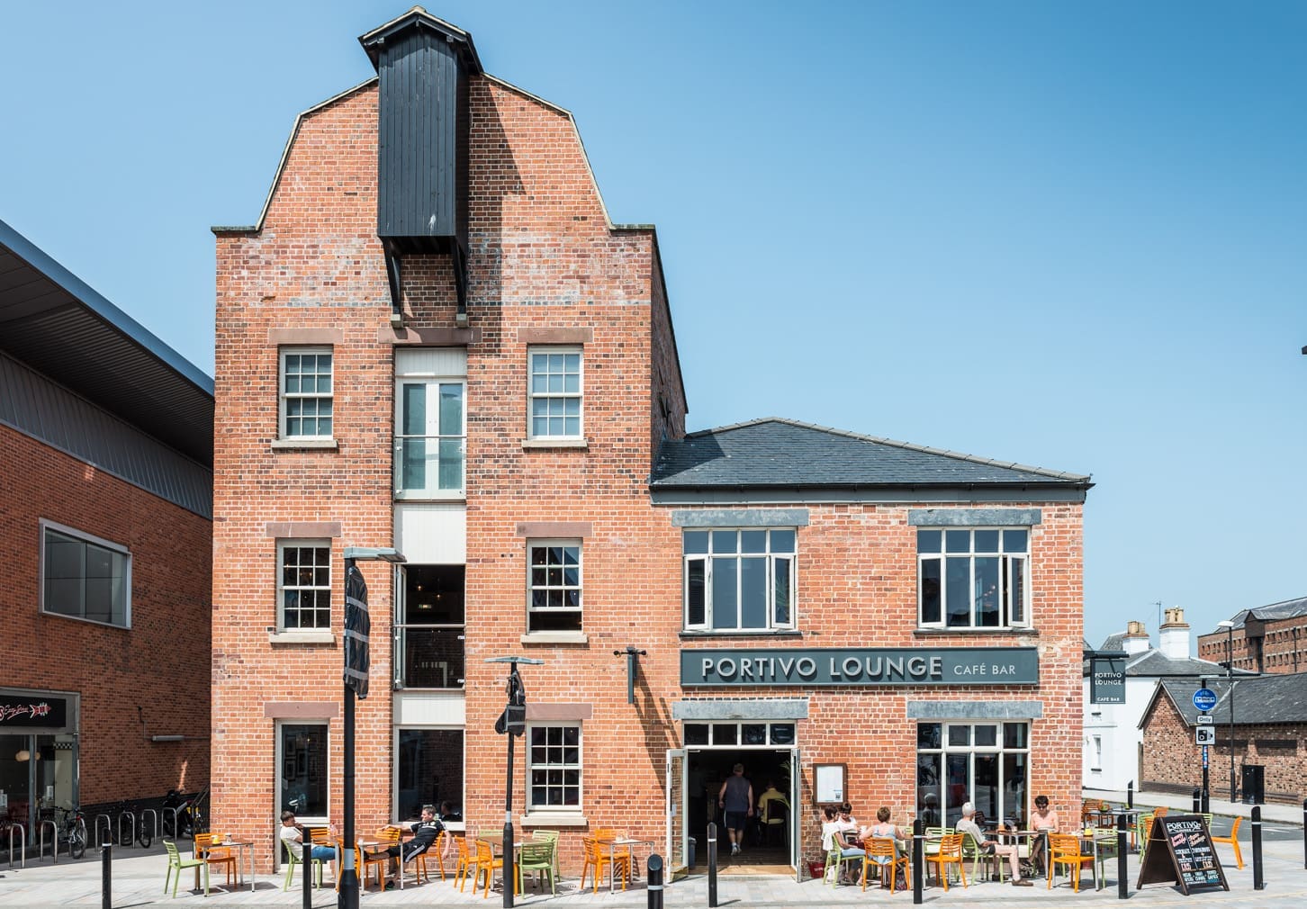 A red-brick building with large windows houses Portivo Lounge Café Bar. People relax at outdoor tables on a sunny day, with the clear blue sky above and nearby buildings framing the view at Portivo.