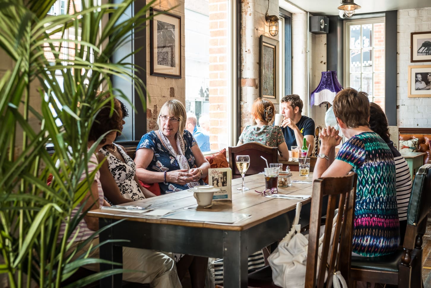 A group of people sit around a wooden table in Portivo, a cozy café, engaged in conversation. Sunlight streams through large windows, framed pictures decorate the brick walls, and a leafy plant is in the foreground.