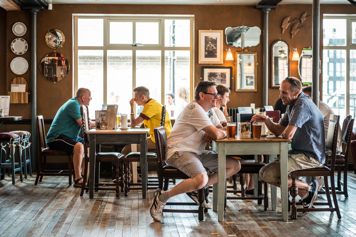 Several men sit at wooden tables in the cozy Portivo pub, chatting and drinking beer. Sunlight streams through large windows, while mirrors and framed photos decorate the inviting walls.