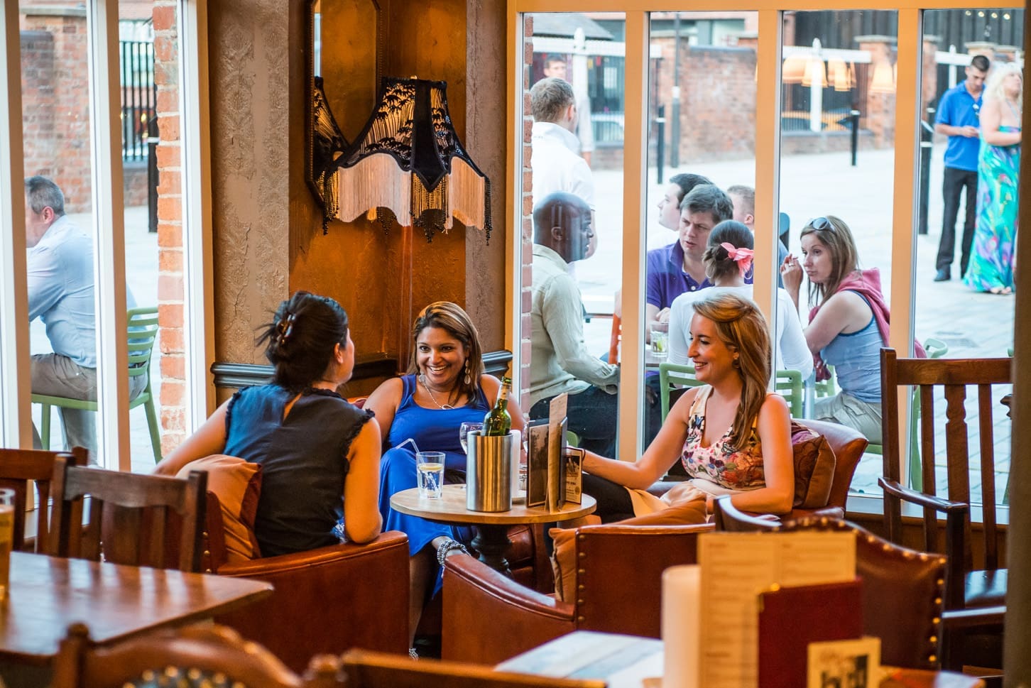 Three women sit at a table inside the cozy Portivo café, smiling and chatting over drinks. Large windows behind them reveal people socializing outside on a patio in a lively, urban setting.