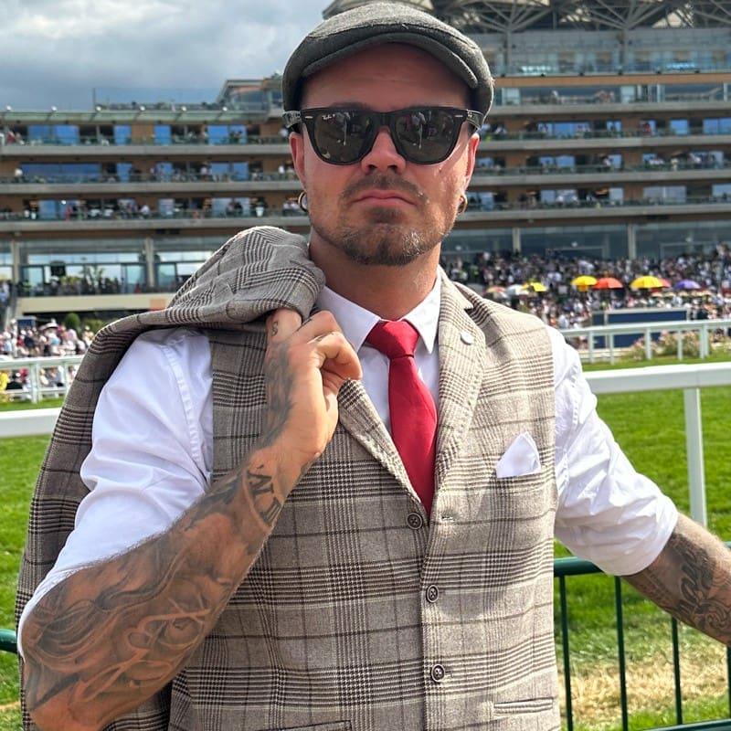 A man wearing sunglasses, a flat cap, and a plaid vest with a red tie stands outdoors at a racetrack, holding his jacket over his shoulder. There are crowds and a grandstand in the background.