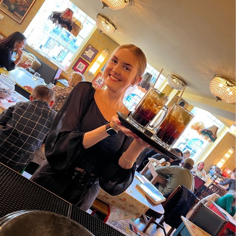 A smiling waitress in a black blouse holds a tray with three tall iced drinks in a busy, warmly lit café filled with seated customers.
