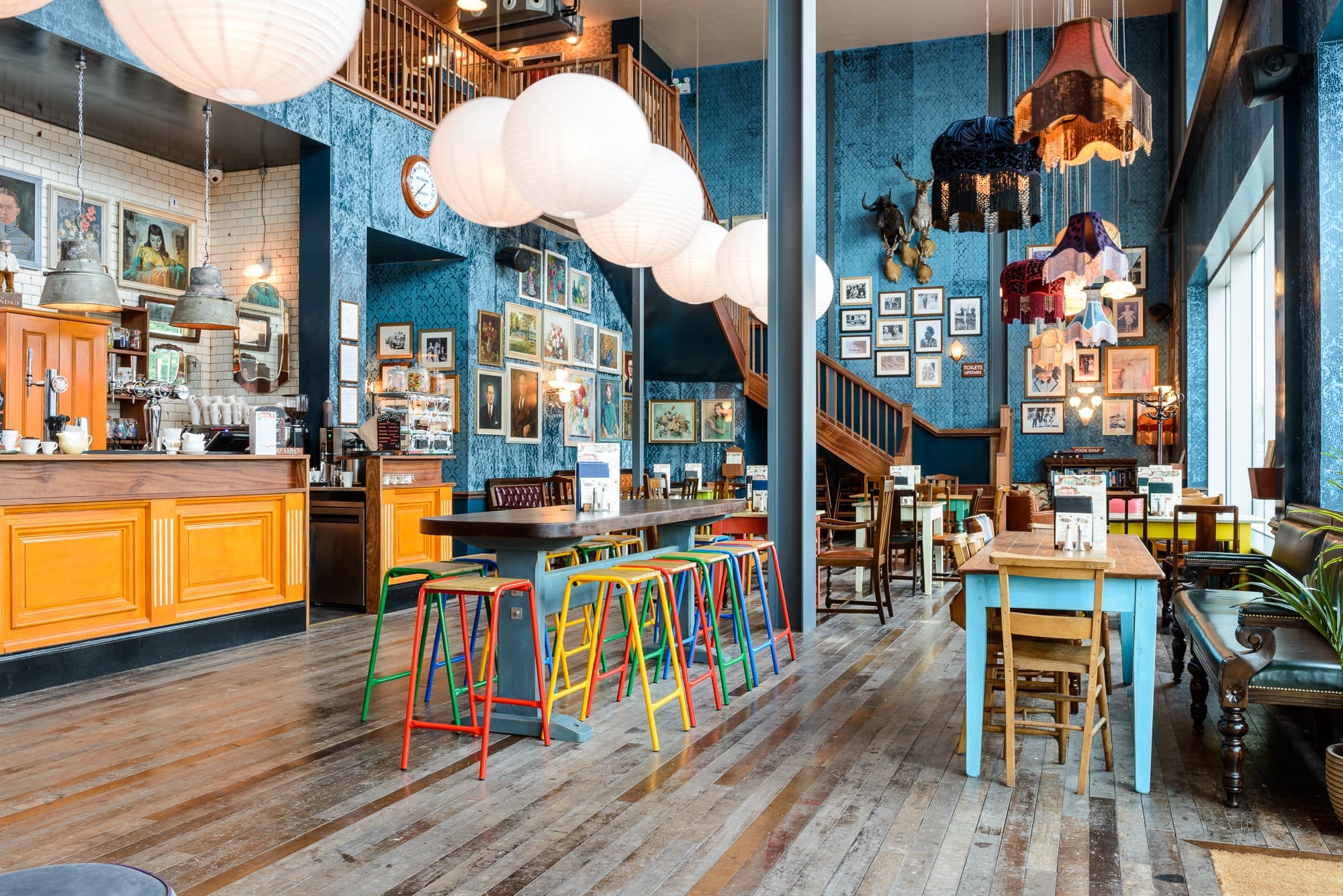 Colorful, eclectic café interior with wooden floors, vibrant Desco furniture, paper lanterns, framed art on blue walls, and a mix of modern and vintage décor. Large windows provide natural light, and a staircase leads to an upper level.