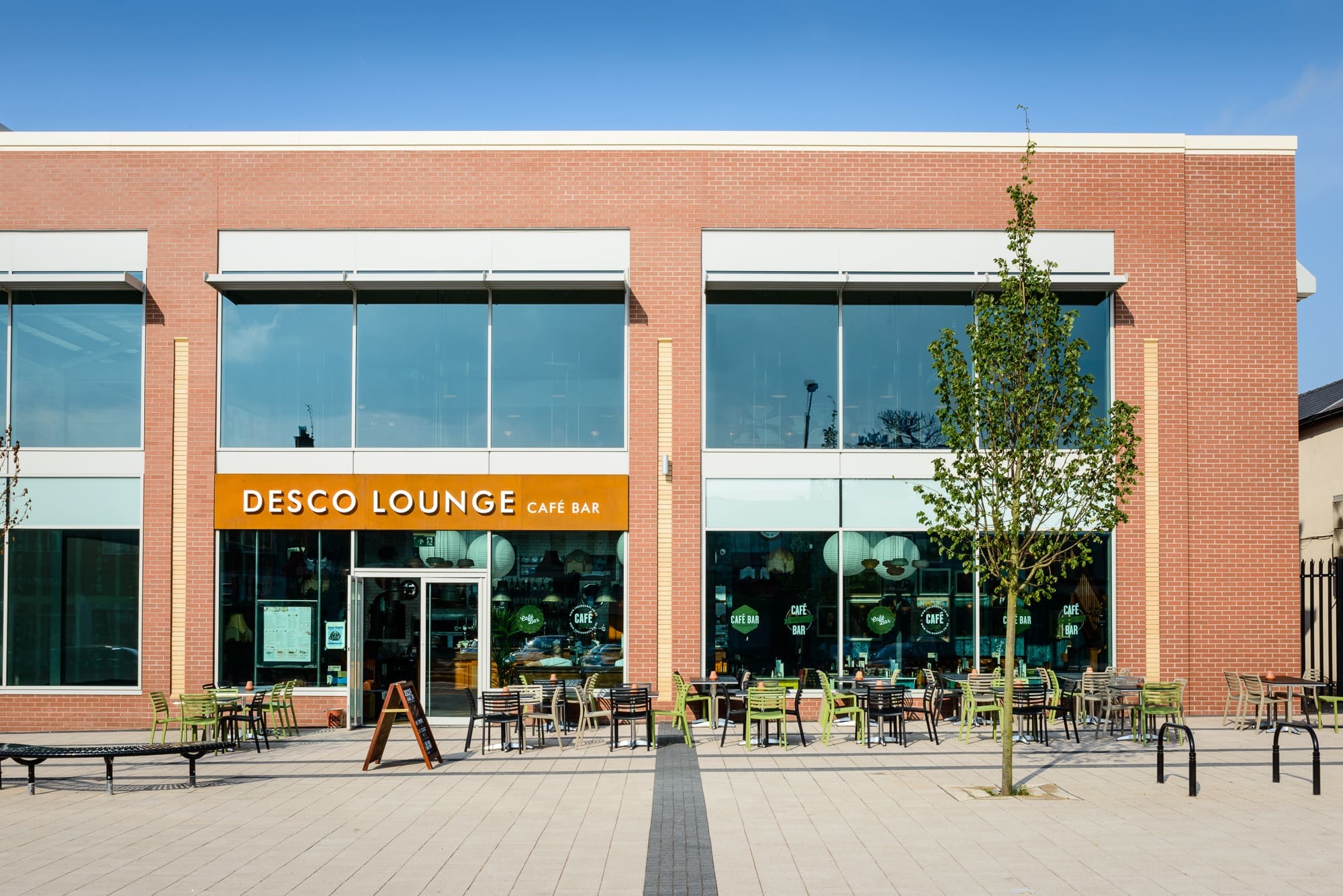 A modern brick building with large windows houses Desco, a vibrant café bar. Outdoor seating with tables and chairs is arranged in front, framed by young trees and a clear blue sky above.