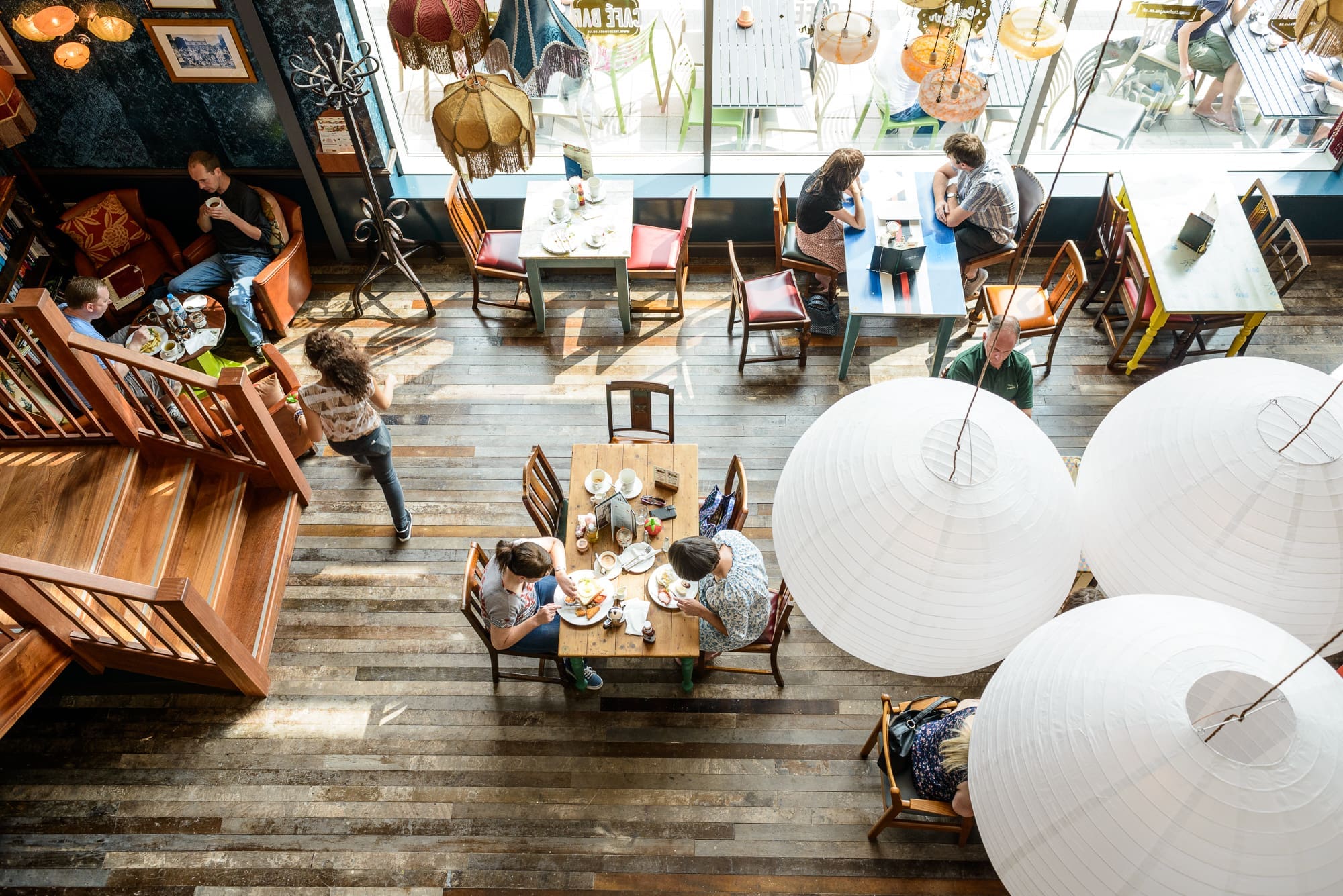 A bright cafe, Desco, seen from above, shows people eating and talking at tables on wooden floors, with large white paper lanterns and sunlight streaming in through expansive windows.