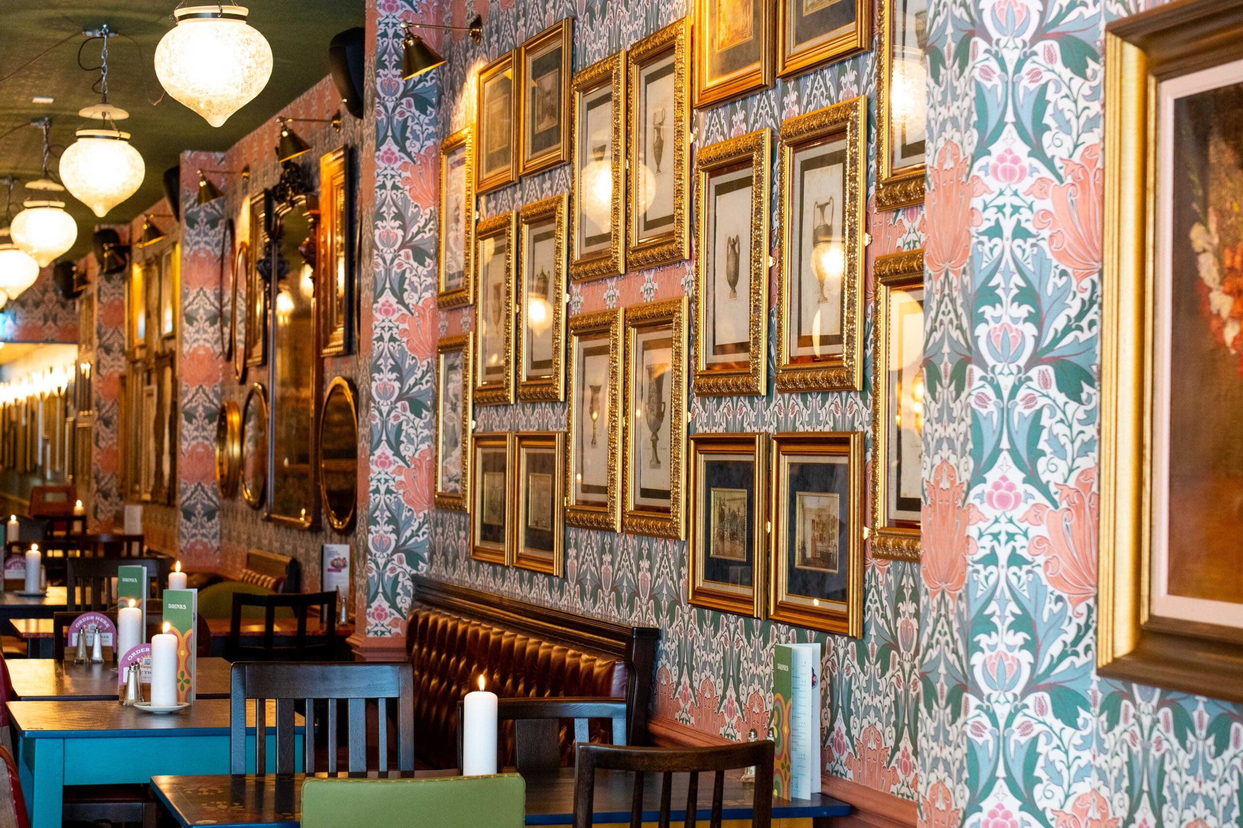 A cozy restaurant with floral wallpaper, framed pictures, vintage light fixtures, and wooden tables set with menus atop smooth granito surfaces. The setting feels warm and inviting.