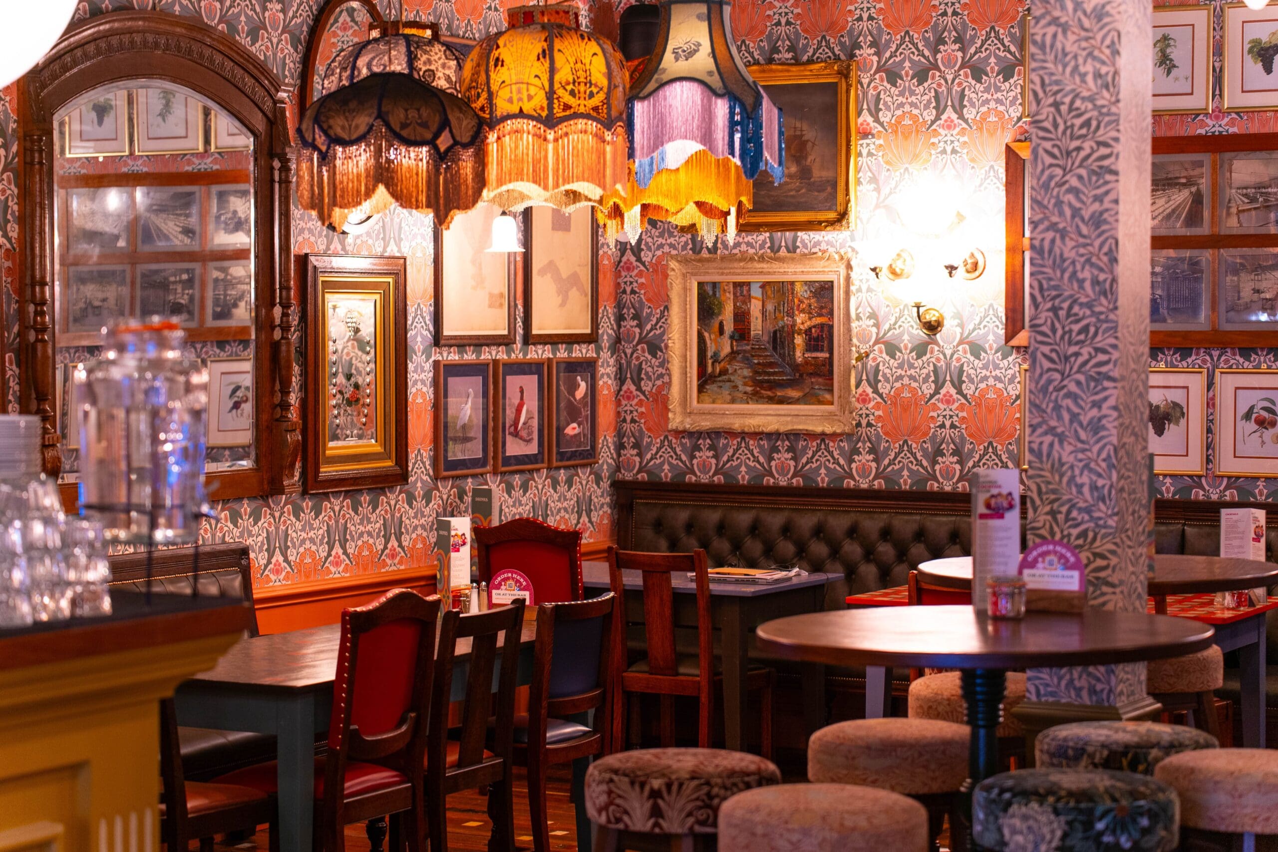 A cozy, vintage-style café with patterned wallpaper, ornate lamps, framed art on the walls, upholstered stools, and wooden tables with menus atop sleek granito surfaces. The warm lighting creates an inviting atmosphere.