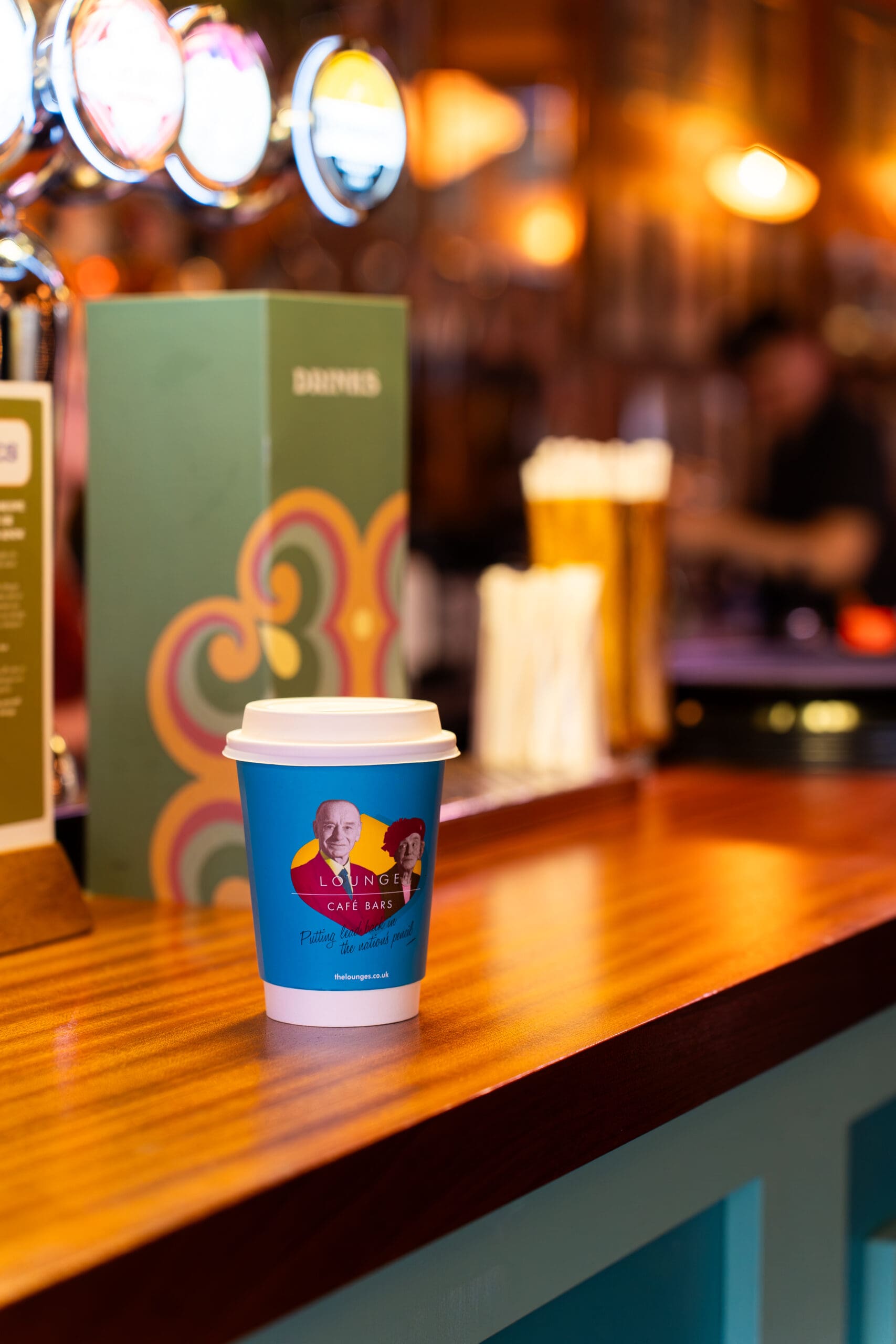 A divino blue takeaway coffee cup with a colorful label sits on a wooden counter in a warmly lit café or bar, with blurred drink taps and a menu in the background.