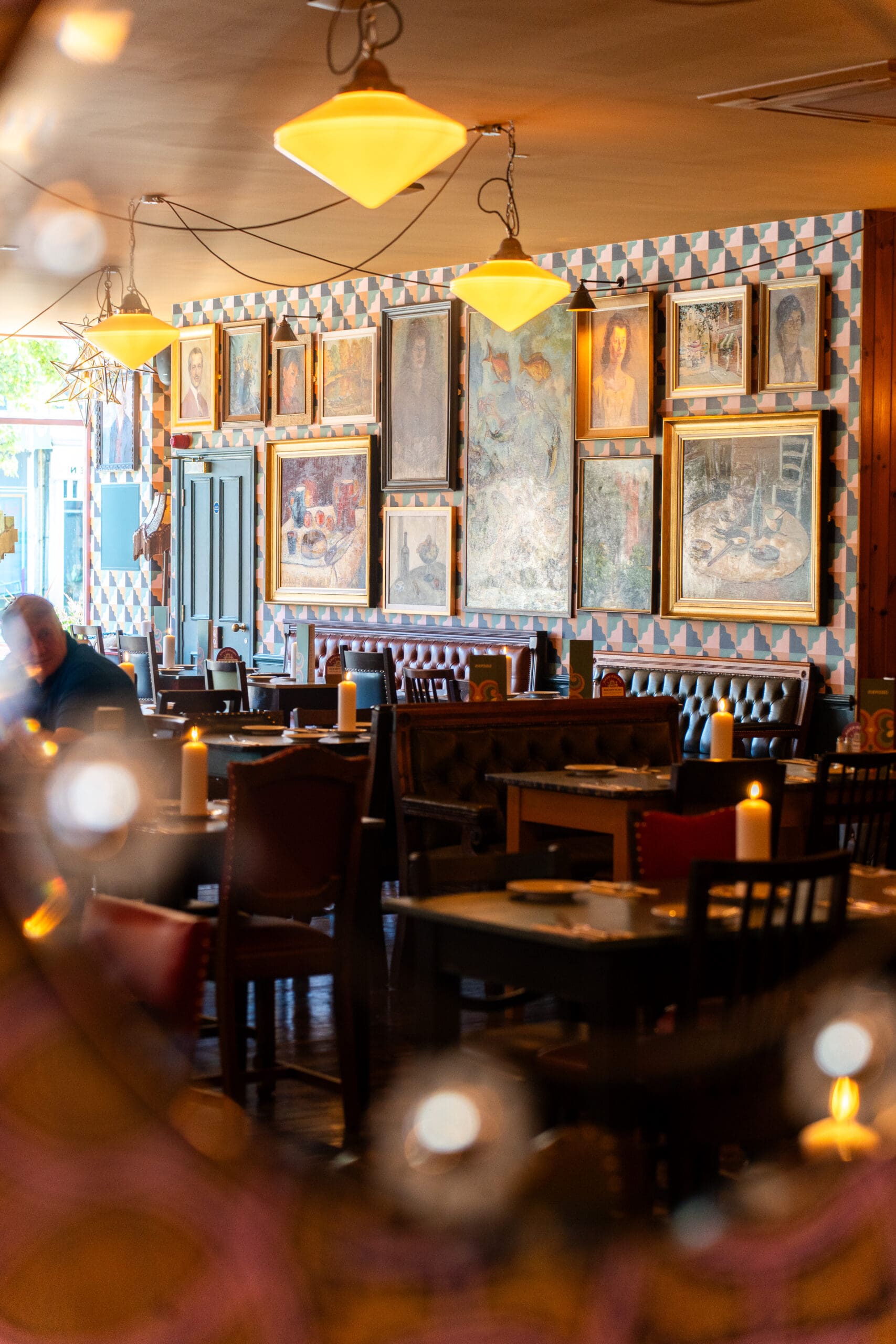 A cozy restaurant interior with wooden tables, red-cushioned chairs, and lit candles. The walls are adorned with divino framed paintings and portraits, creating a warm, artistic atmosphere where a few people are seated at the tables.