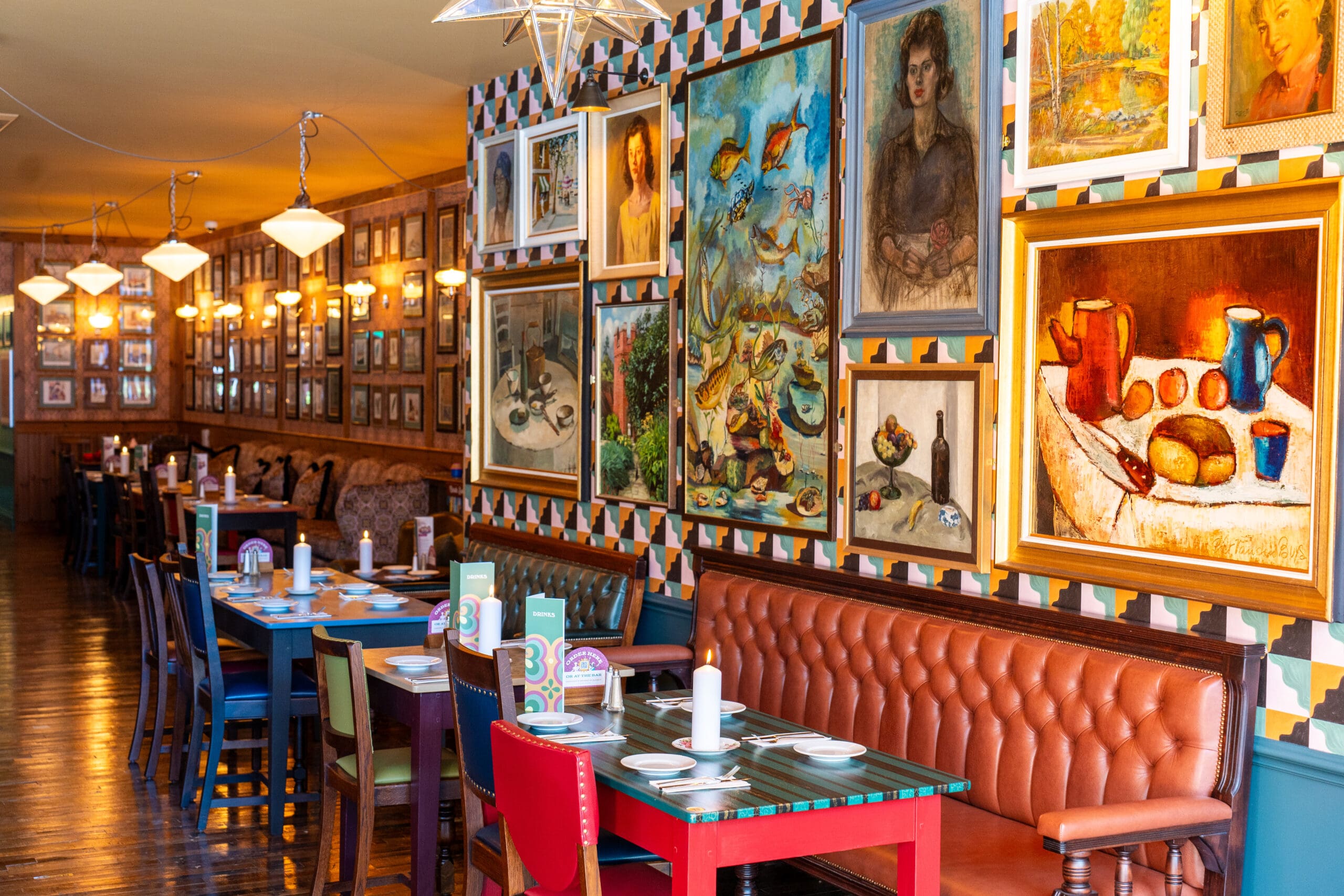 Vibrant restaurant interior with colorful chairs, patterned walls, and eclectic artwork—including portraits, still lifes, and abstract paintings—creates a divino atmosphere. Tables are set with menus and napkins along a tufted leather bench.
