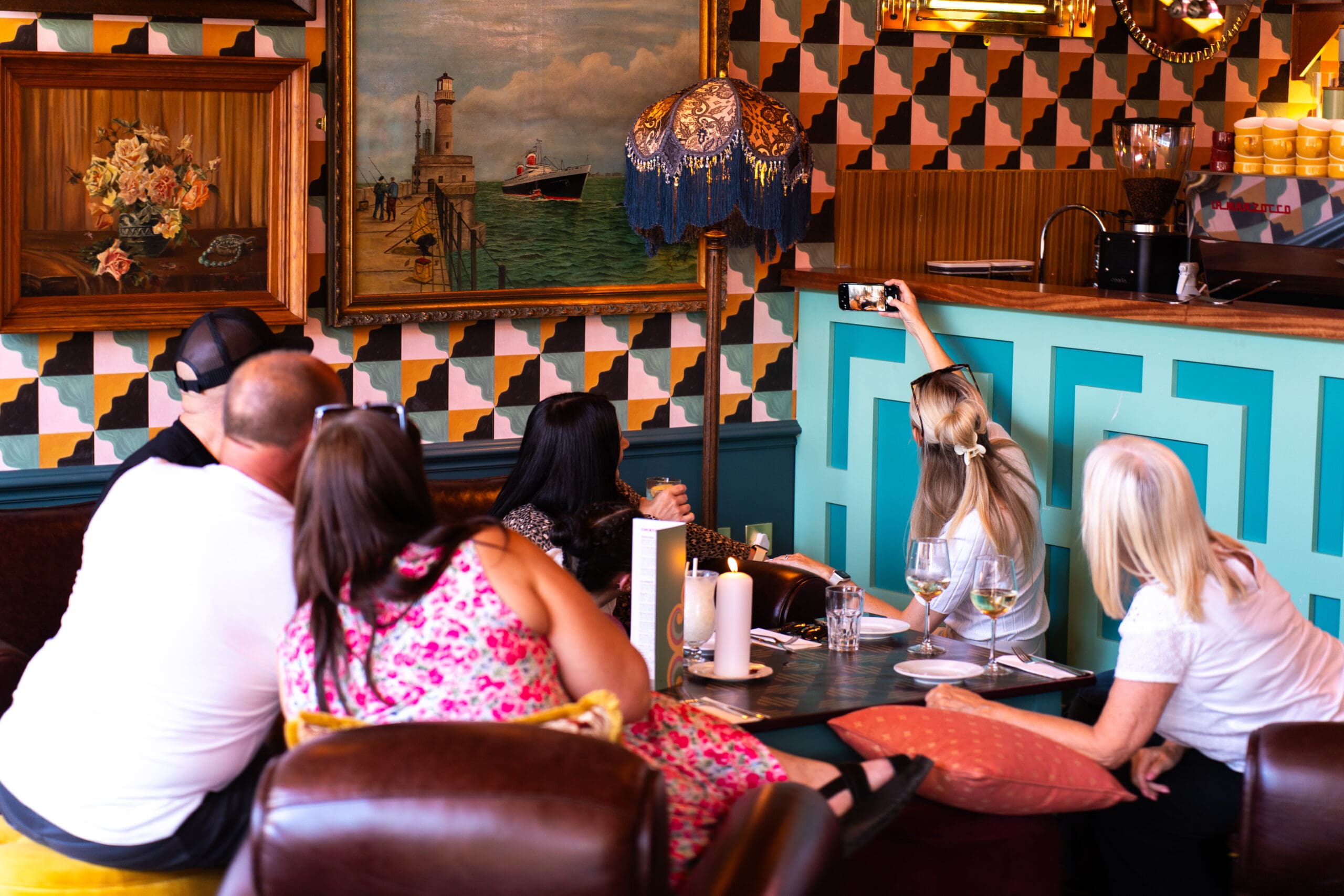 Four people sit around a table in a colorful, retro-styled café. One woman stands to take a divino selfie of the group. Drinks and menus are on the table, and vintage art decorates the walls.
