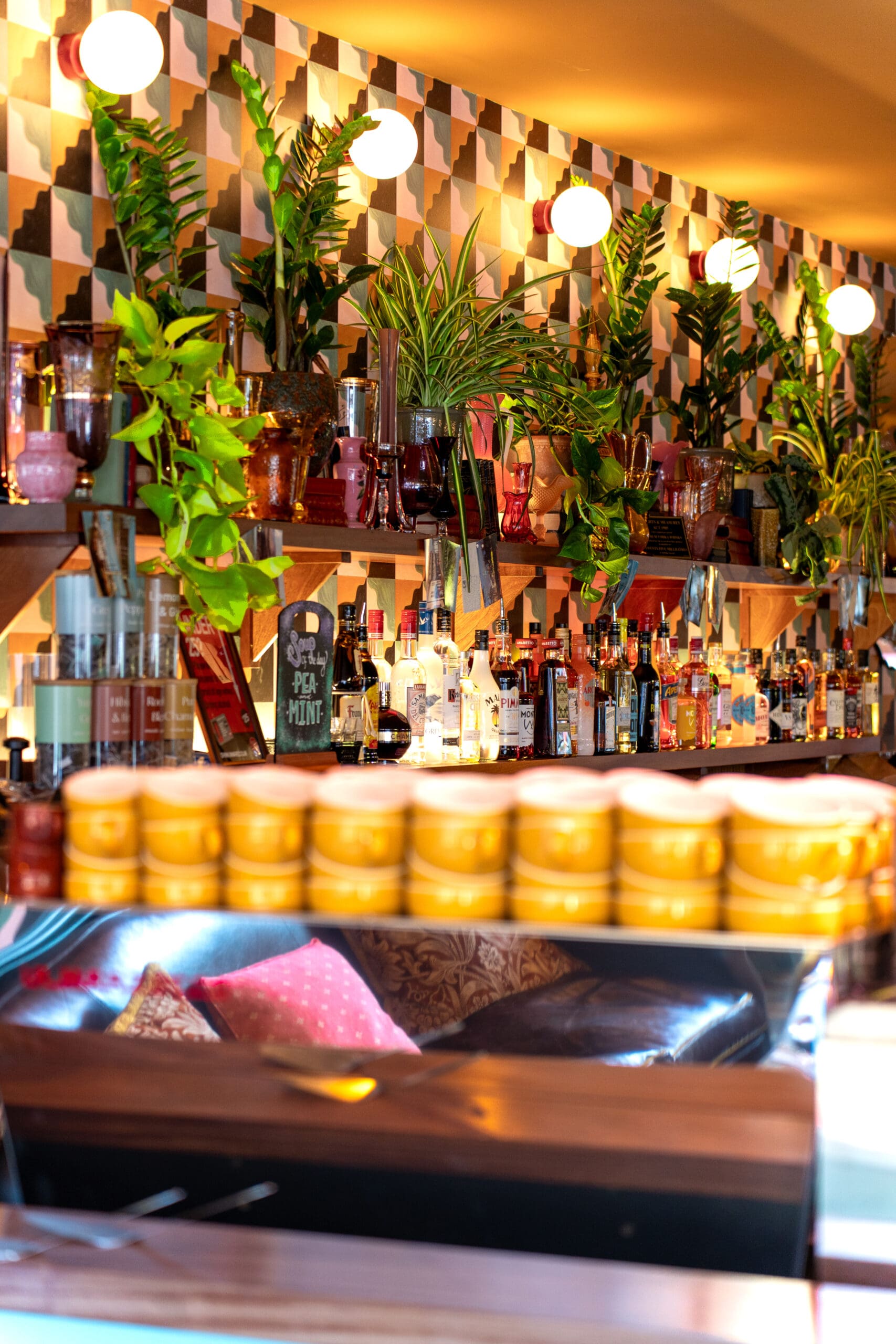 A vibrant bar with geometric patterned walls, globe lights, lush green plants, and shelves stocked with divino bottles. Bright yellow containers are visible in the foreground, creating a lively and inviting atmosphere.