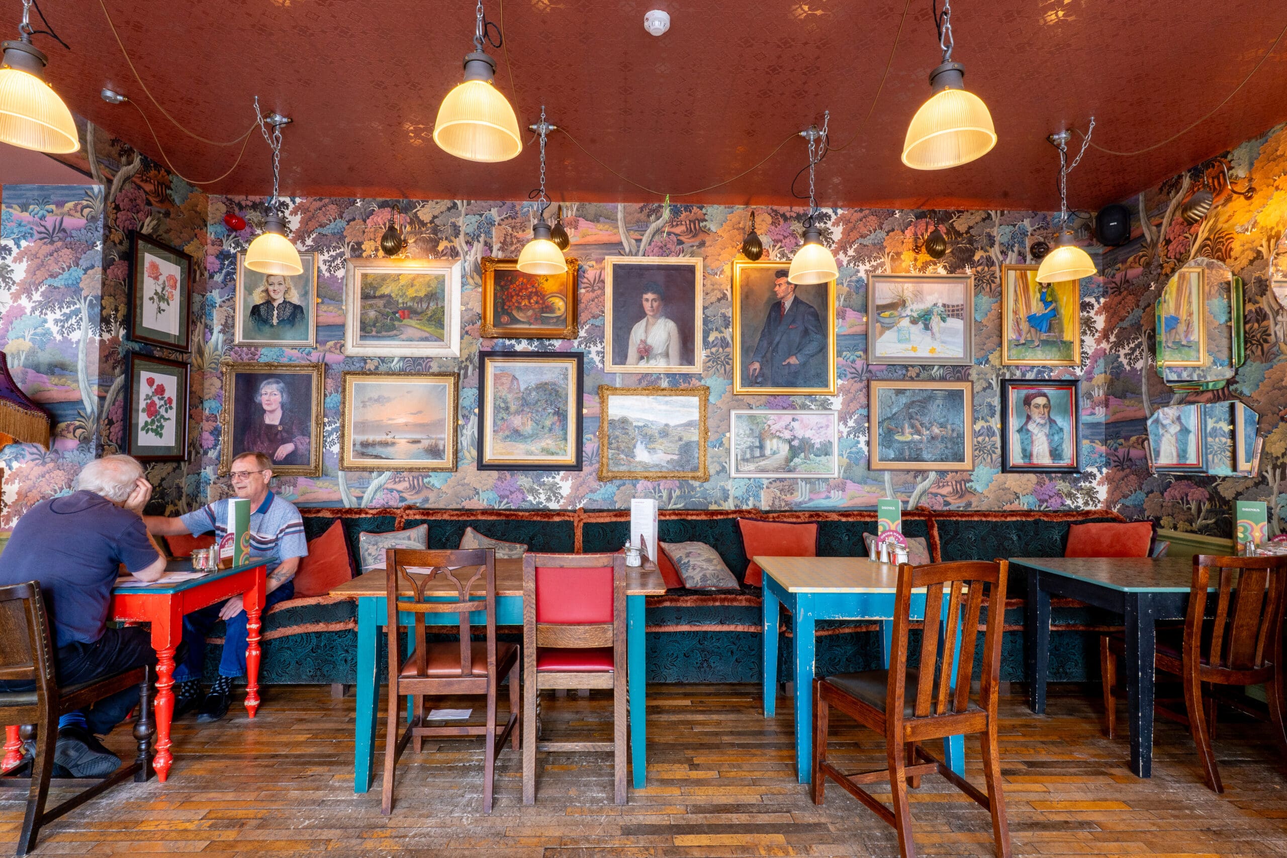 Colorful café interior in Porto with eclectic tables and chairs, pendant lights above, and walls covered in framed portraits and paintings. Two people converse at a table on the left, with empty tables in the foreground.