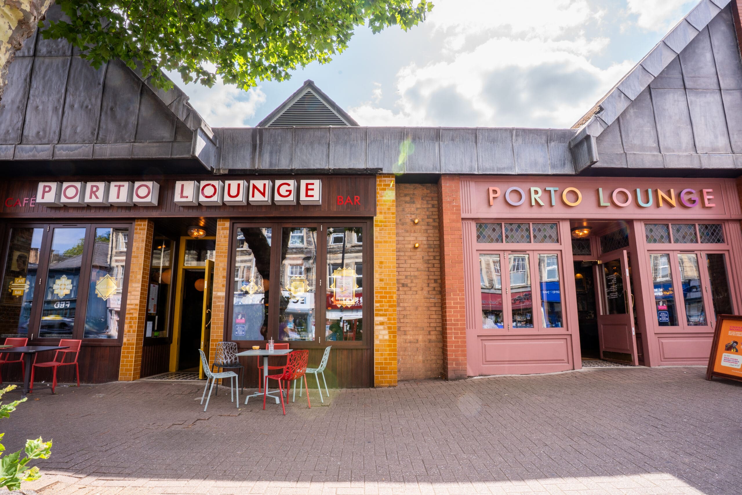 Exterior of Porto Lounge café and bar in Porto, showing two entrances, outdoor tables and chairs, and large red and pink signage under a partly cloudy sky.