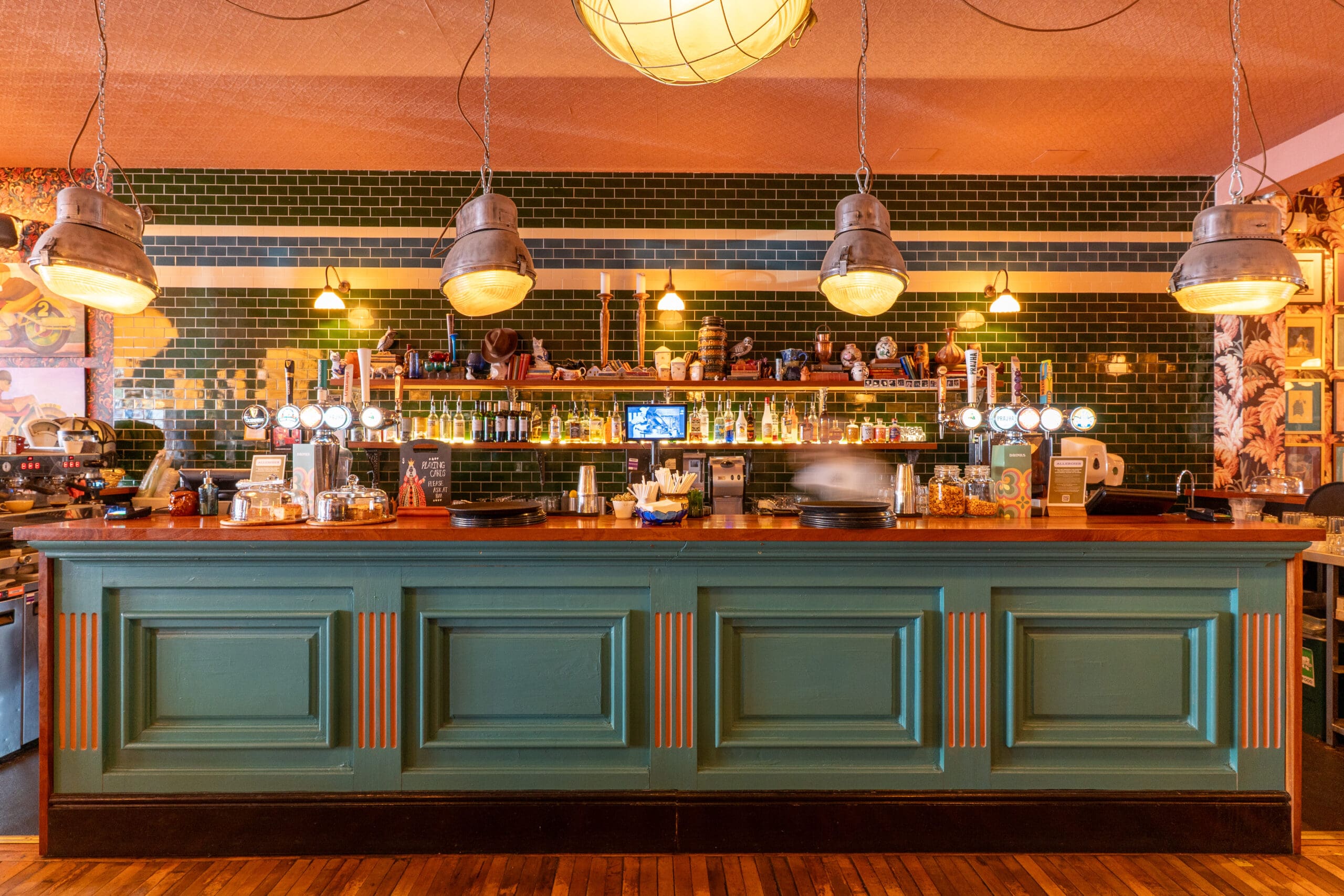 A stylish Porto bar with a green paneled front, green tiled wall, hanging industrial lights, bottles, glasses, and beer taps arranged neatly on shelves behind the counter.