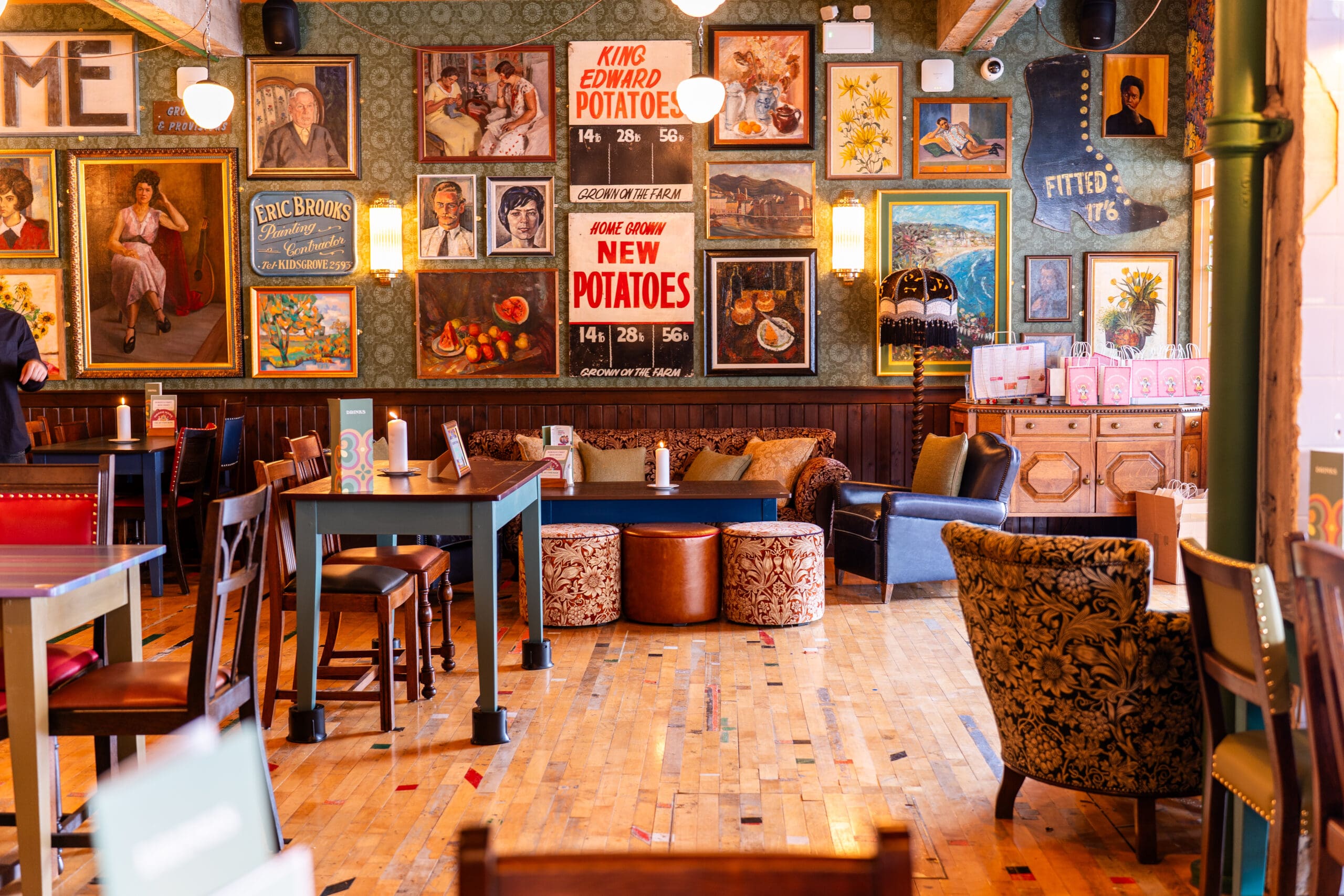 A cozy, vintage-style cafe with eclectic chairs, wooden tables, and marinero-themed walls decorated with framed art and retro posters. Warm lighting and patterned wallpaper create an inviting atmosphere.