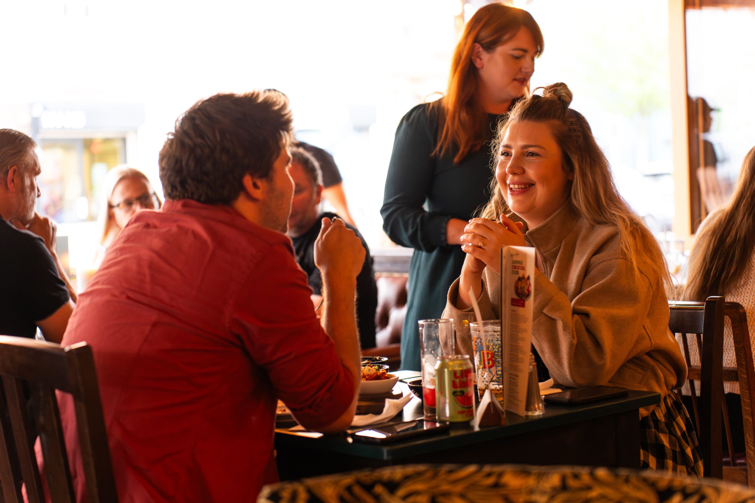 A group of people sit at a restaurant table, talking and smiling. A woman laughs while looking at a man across from her. Another woman stands nearby, listening and smiling. Bottles, menus, and a marinero hat are on the table.