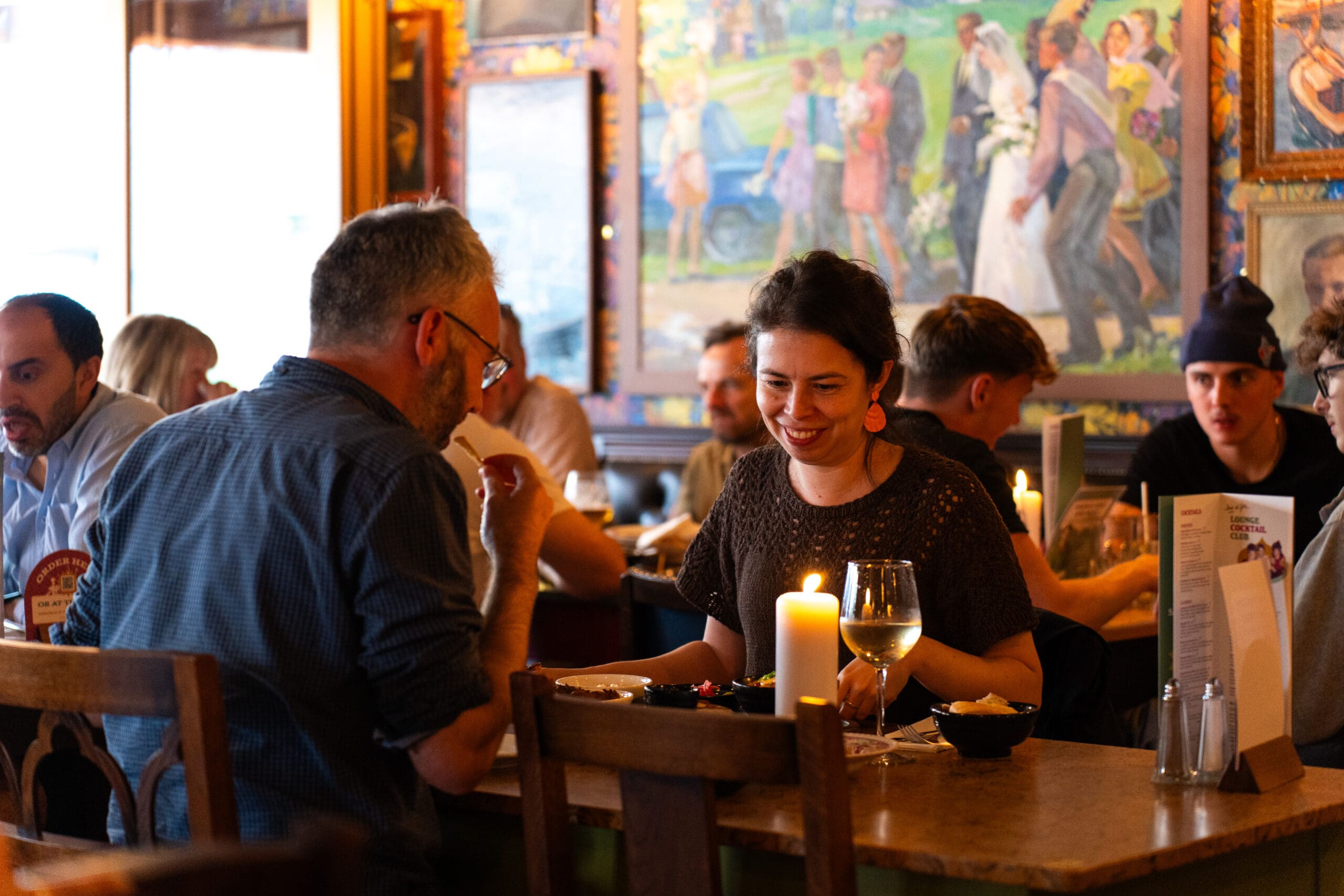 A woman and a man dine together at a warmly lit rubino-hued restaurant, smiling and talking. Other people are seated nearby, candles and drinks on the table, as colorful art decorates the walls in the background.