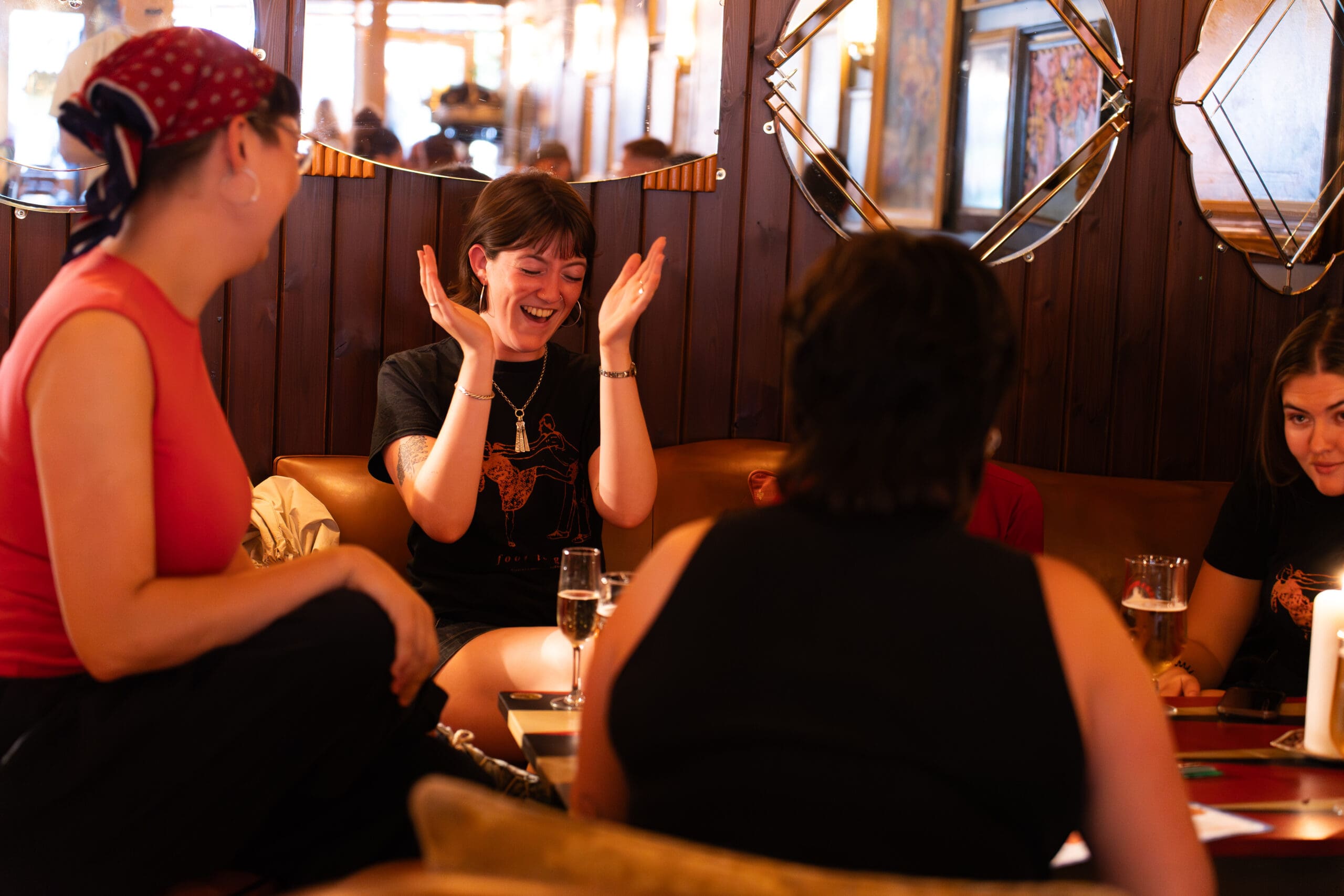 A group of women sit around a table in a cozy, warmly lit room, laughing and enjoying drinks. One woman in the center is clapping her hands and smiling as her friends cheer with marinero cocktails, surrounded by lively conversation.